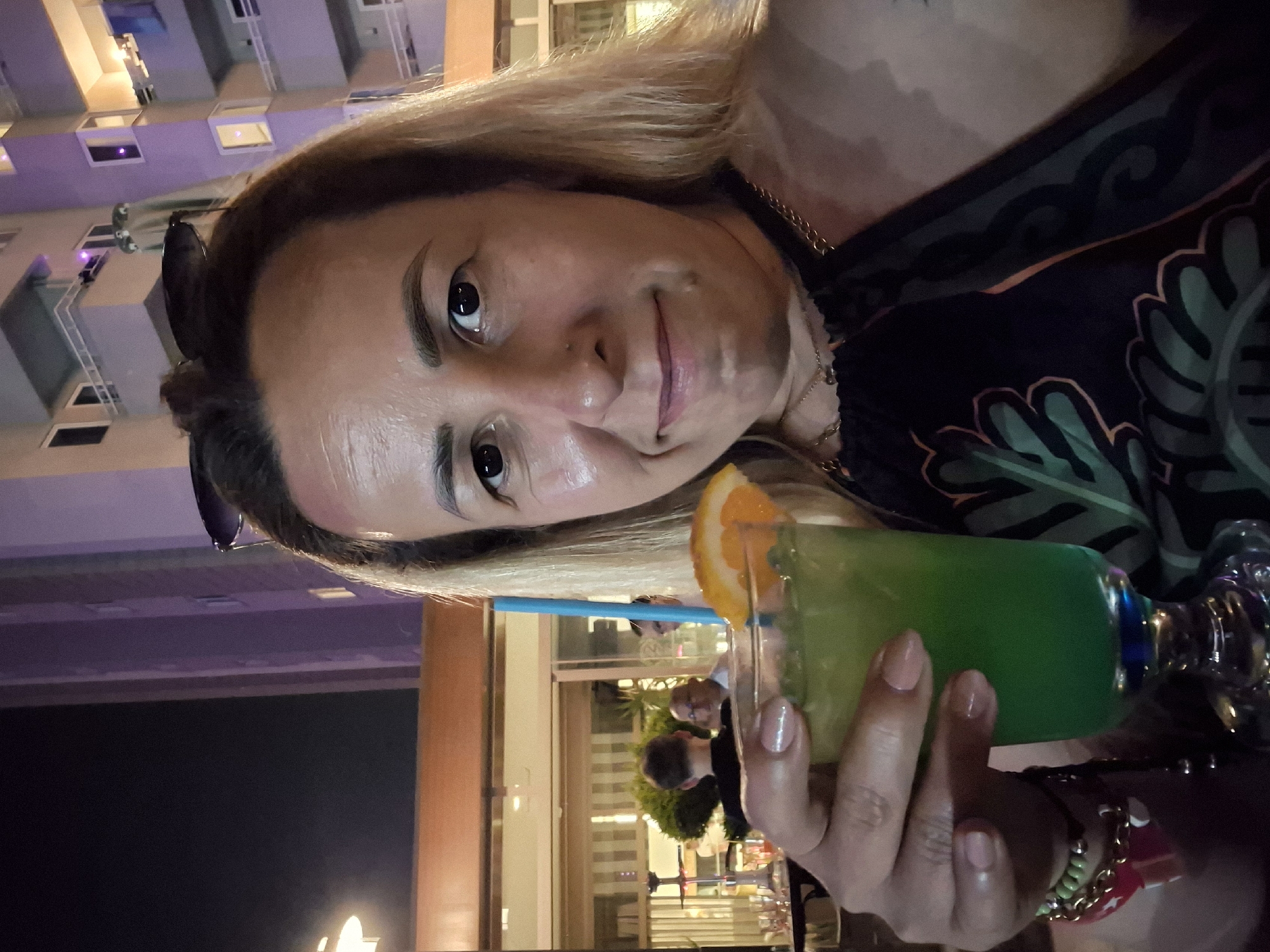A woman holds a green cocktail with an orange slice garnish. She has blonde hair and is wearing a dark patterned top. She is holding the drink with her right hand, which has light pink nail polish and several bracelets on her wrist. In the background, a building with purple and white accents is visible, with lights illuminating some of its windows. The scene appears to be outdoors at night, as indicated by the darkness and artificial lighting. The woman's face is partially visible, and she has makeup on, including eyeliner and lipstick. There are other people in the background, slightly out of focus, suggesting a public setting like a restaurant or bar.
