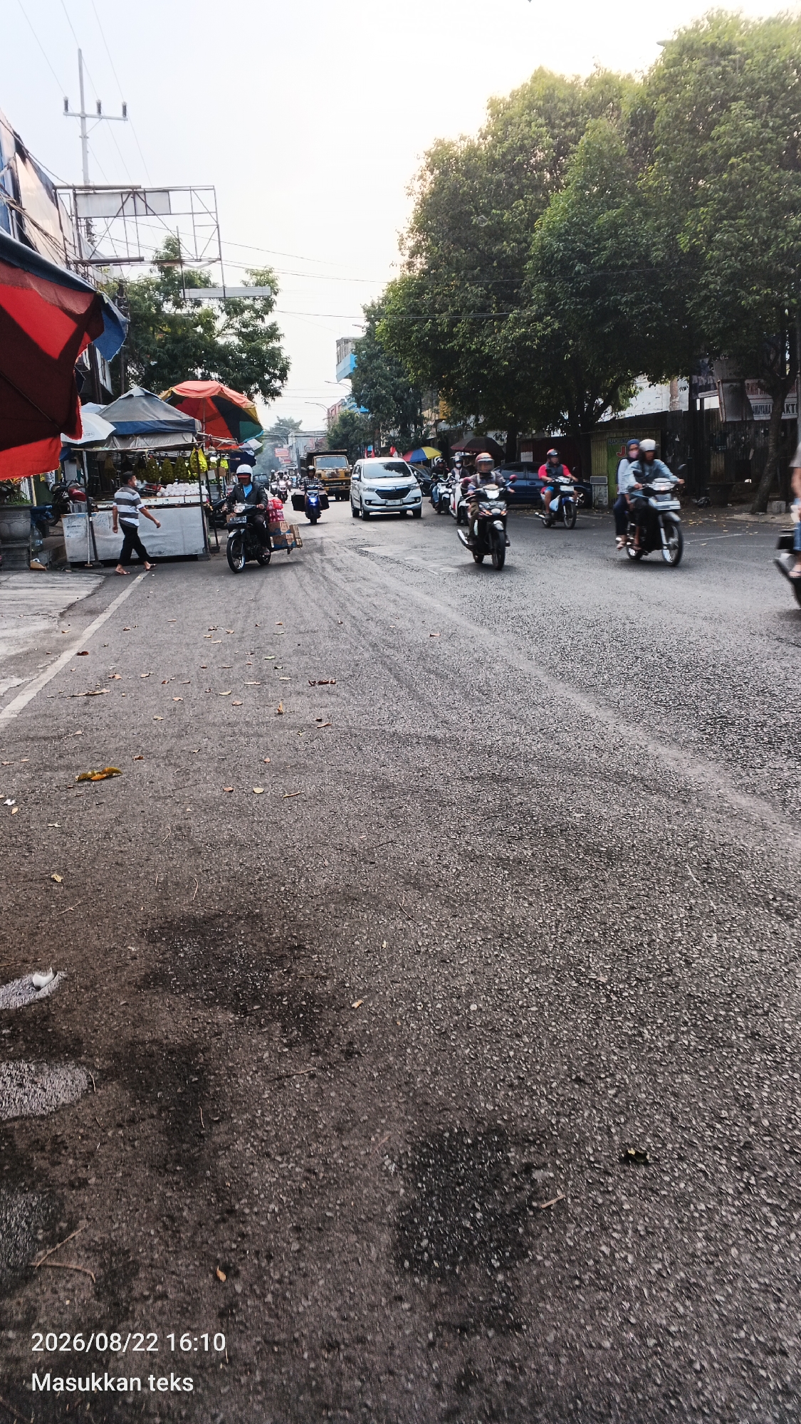 The image depicts a street scene in Malang, Indonesia, with a road filled with various vehicles and pedestrians. On the left side of the road, there are stalls with colorful awnings, likely part of a market or roadside vendors. Several people are visible, some walking and others on motorcycles. A white car is in the center of the road, surrounded by motorcycles. Trees line the right side of the street, and buildings are visible in the background. The sky is bright, suggesting it is daytime, possibly late afternoon due to the warm light. Debris, including leaves and what appear to be small branches, is scattered on the asphalt road. The timestamp "2026/08/22 16:10" and "Masukkan teks" are superimposed on the bottom left corner of the image.