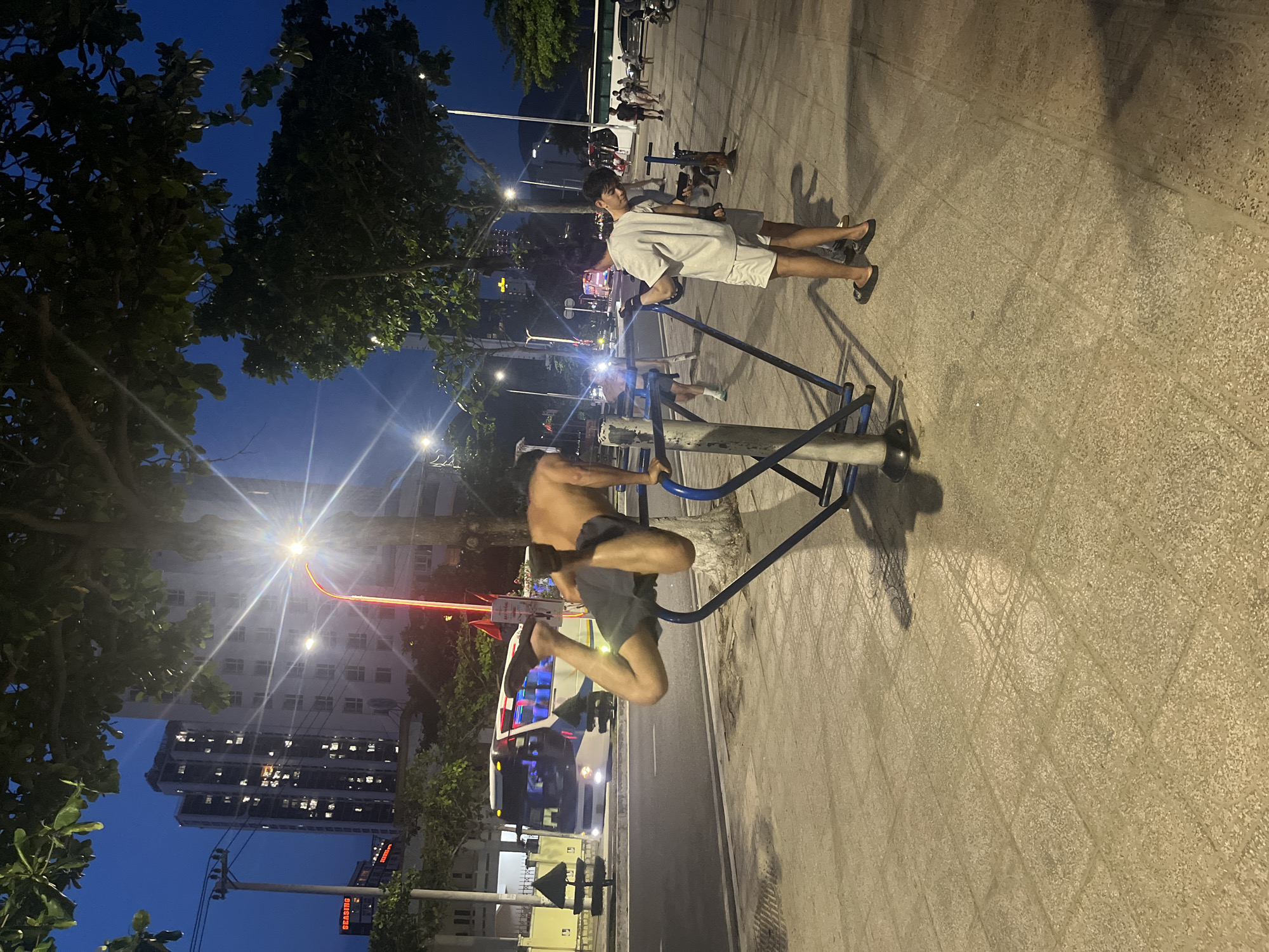 A nighttime scene presents several individuals and objects along a city street in Unknown, Unknown. A man is positioned on a blue fitness machine and seems to be leaping off, displaying his bare chest and dark shorts. Two people are positioned on another workout machine nearby. The environment includes a variety of elements: a city skyline of tall buildings in the distance, trees lining the streets, street lights illuminating the scene, and various parked bicycles. The road itself is a smooth asphalt surface, and the walkway has patterned concrete tiles. The sky is a deep blue, suggesting it's either early evening or late night.