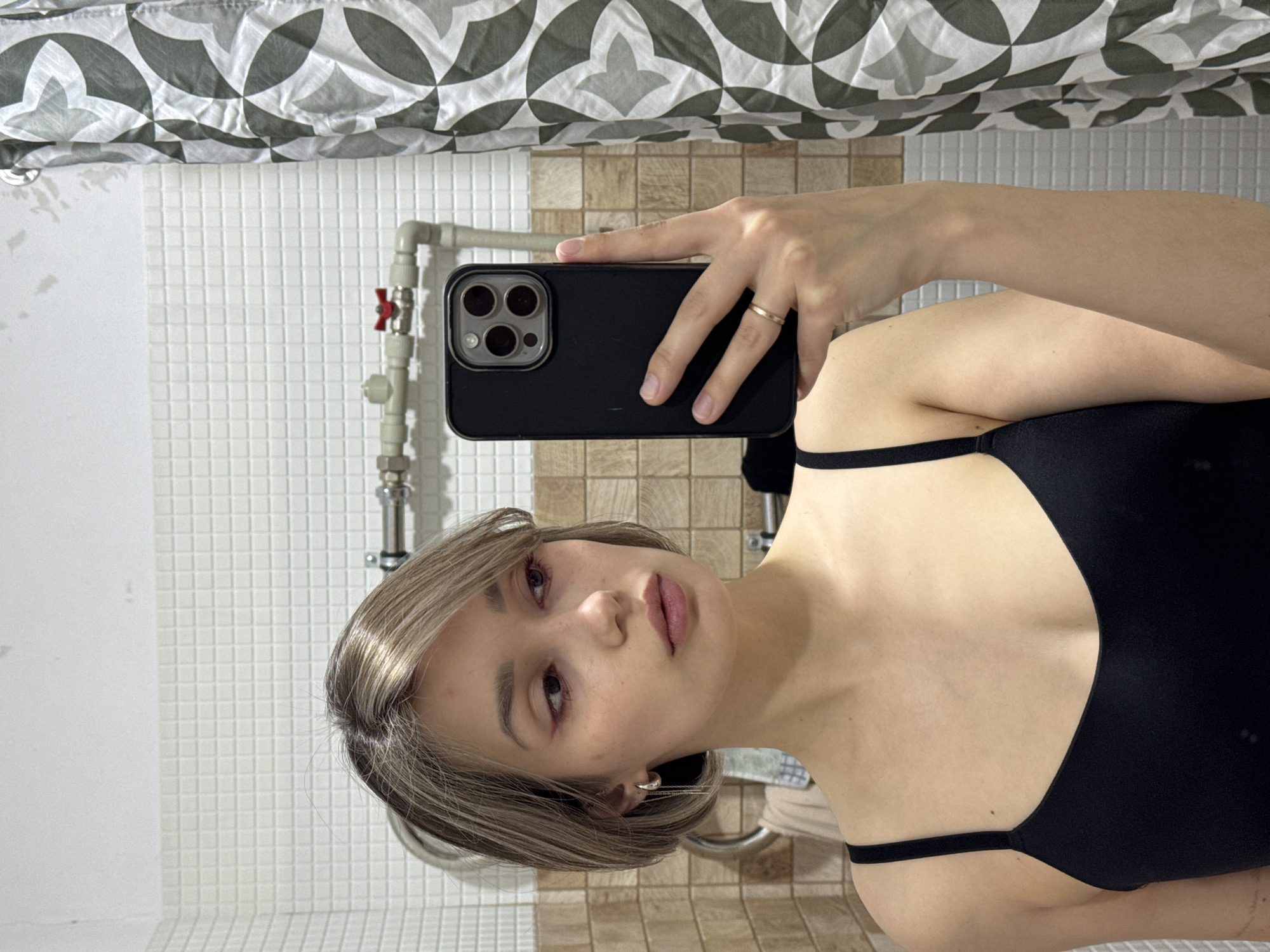 A woman is taking a selfie in a bathroom. She is holding a black smartphone with multiple camera lenses in her right hand. The woman has short, light brown hair and is wearing a black top with thin straps. The bathroom walls are tiled; the wall behind the woman features small white square tiles, while the wall to her right has larger brown-toned square tiles. A shower curtain with a grey and white geometric pattern is visible at the top of the frame. A showerhead and some pipes are visible against the white tiled wall. The location is Unknown, Unknown.