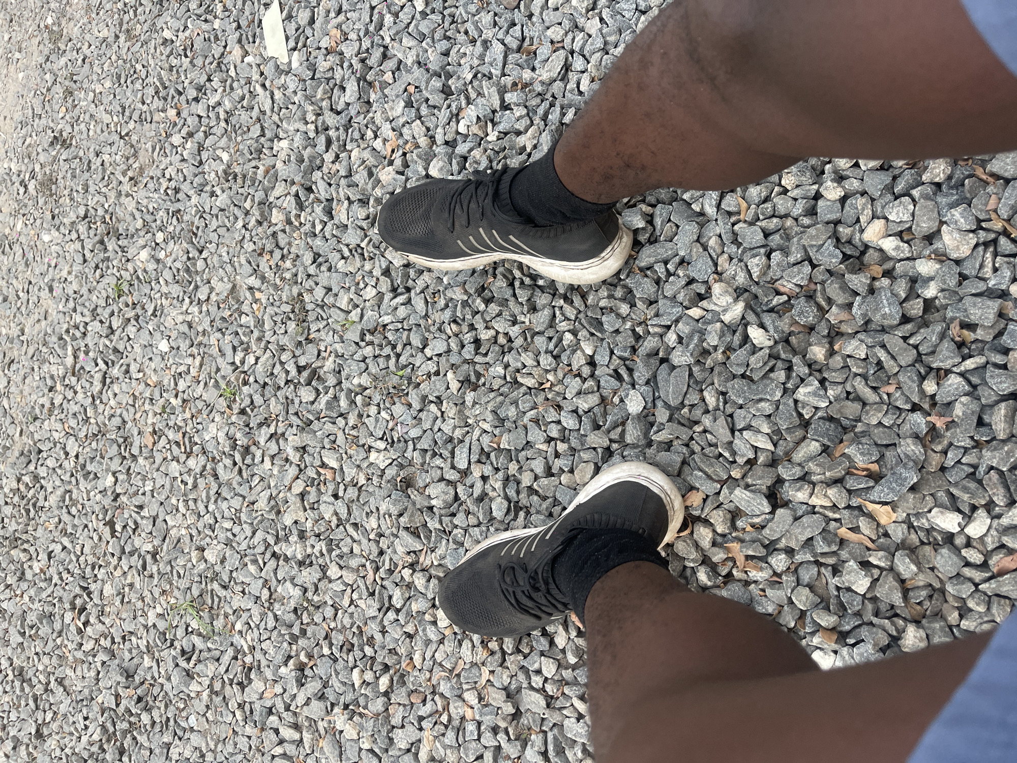 A person stands on a surface covered in small gray rocks. The person is wearing black athletic shoes with white soles and black socks. The view is from above, looking down at the person's legs and feet. A blue garment is visible on the right. The location is Unknown, Unknown.