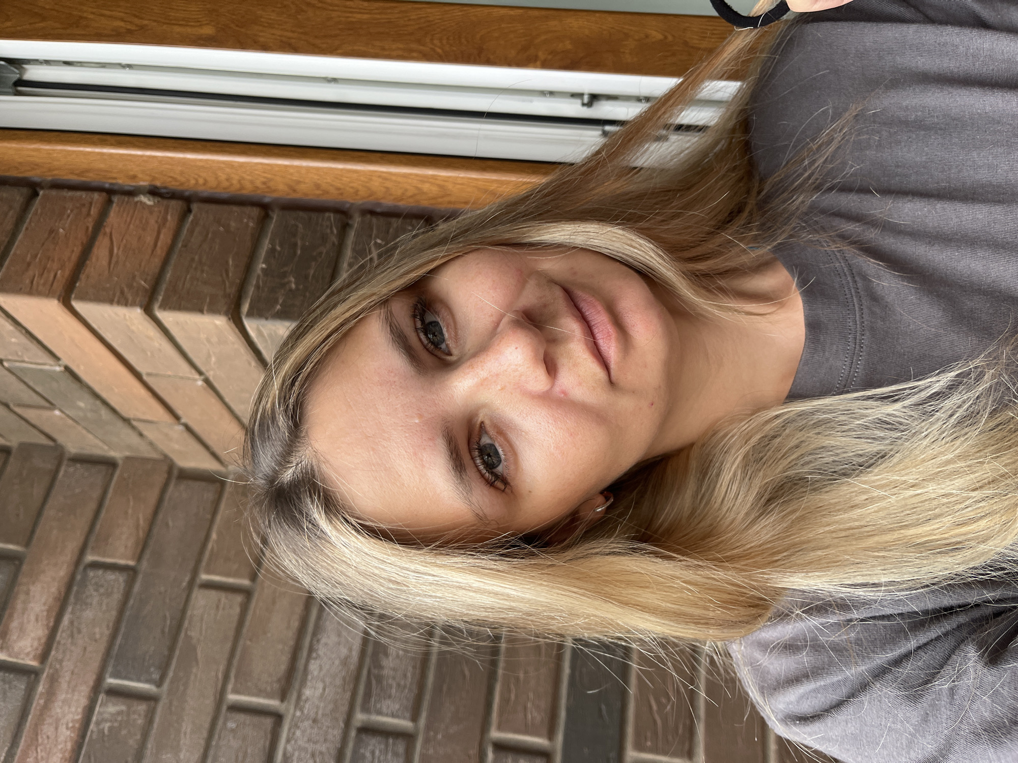 A young woman with blonde hair and blue eyes is pictured in front of a brick wall and a wooden door. She is wearing a gray t-shirt. Her hair is slightly windswept, and she is holding a black object in her right hand, partially visible at the edge of the frame. The lighting suggests it is daytime. The background brick wall appears to be an exterior wall of a building, possibly in Pionersky, Russia, given the context.
