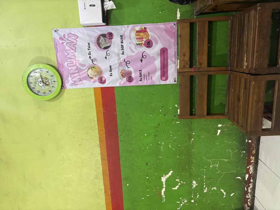 The image depicts an interior scene, likely a beverage stall or eatery, located at Perum Orchid Regency A15 in Sidodadi, Indonesia. The wall is horizontally divided into three sections: a lime green upper portion, a central band featuring distinct orange and red stripes, and a lower, darker green section exhibiting extensive wear, including visible cracks, peeling paint, and underlying white and red layers.
Mounted on the lime green wall is a round, light green clock with a cartoon frog character (Keroppi) on its face. Below the clock and extending into the lower wall section, a vertical white and pink banner, labeled "MINUMAN" (Drinks), is displayed. This banner advertises "Es Teler," "Es Oyen," "Es SOP BUAH," and "Es JUICE" (with options including Alpukat, Melon, Semangka, Apel), each priced at 10,000 Indonesian Rupiah ("10k") and depicted with accompanying images. The banner also specifies the location as "Perum Orchid Regency A15." To the right of the banner, a white wall-mounted tissue dispenser, branded "SUBRON," is affixed, with a white tissue sheet partially visible.
In the foreground, two dark brown wooden chairs with slatted backs and seats are positioned side-by-side on a white tiled floor, facing away from the viewer. A portion of a third wooden object is visible on the far right edge of the frame. No individuals are present in the scene.