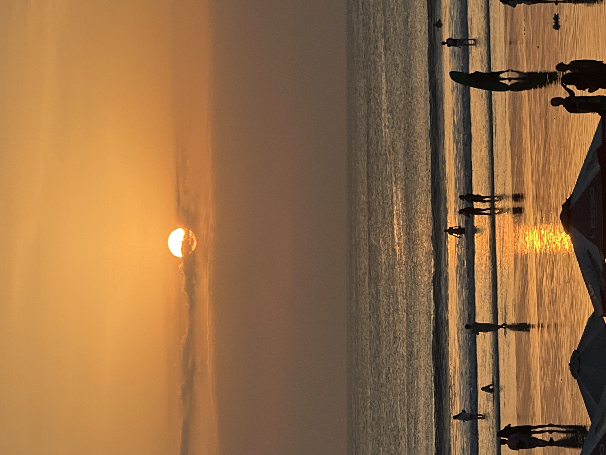 A serene sunset unfolds over the ocean at Kuta Beach, Bali, Indonesia. The sun, a bright orb partially obscured by thin clouds, casts a warm, golden glow across the sky and the wet sand. Gentle waves lap the shore, reflecting the vibrant hues of the sunset. Several people are visible on the beach and in the shallow water, appearing as silhouettes against the bright backdrop. Some individuals are standing, others are walking, and a few appear to be wading in the sea. One person is holding a surfboard. The scene is tranquil, capturing a moment of leisure as the day ends.