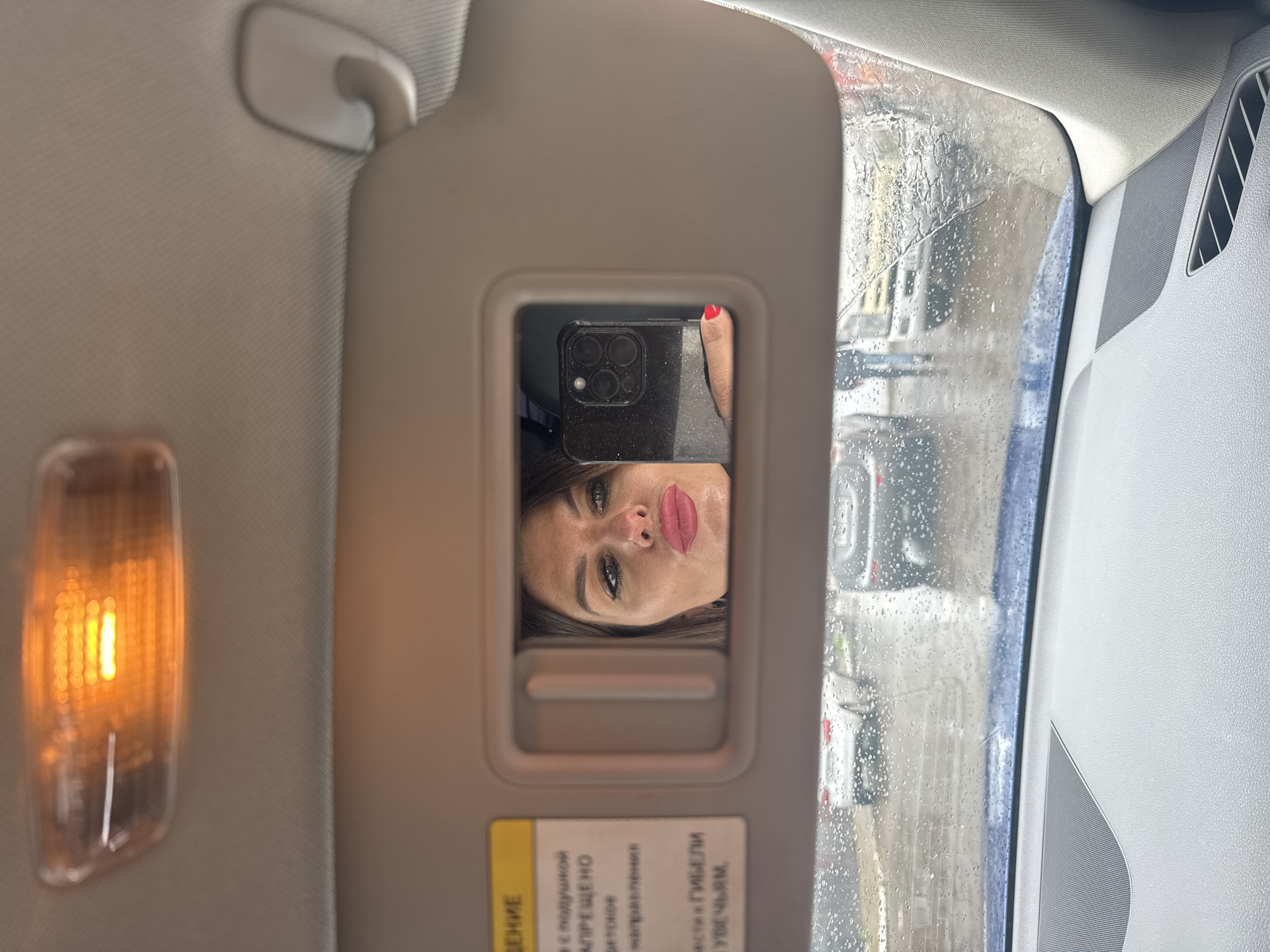 The image captures a selfie taken from inside a car, focusing on the reflection in a small mirror embedded in the passenger-side sun visor. The main subject is a woman's face, seen reflected upside down and slightly angled, distinguished by vibrant red lipstick and prominent eye makeup. Her hand, with a red painted fingernail, holds a black smartphone (identifiable as an iPhone due to its triple camera array) that is being used to take the picture.
The setting is the interior of a car. The sun visor is flipped down, revealing the mirror. To the left, an illuminated overhead light casts a warm, orange glow. The car's ceiling, side pillar, and an air vent or speaker grille are visible on the right.
Beyond the car's interior, the external environment is visible through a heavily rain-splattered window, indicating wet weather and an overcast day. The blurred exterior view shows indistinct shapes of buildings and other vehicles, suggesting an urban or suburban location in Chaikovsky, Russia.
On the sun visor, below the mirror, a yellow warning label with Cyrillic text is present. Partially visible words include "ВНИМАНИЕ" (ATTENTION/WARNING), "ПОДУШКОЙ" (airbag), and "ПРЕЩЕНО" (forbidden/prohibited), indicating a standard automotive safety warning, likely related to airbags. The woman's expression is neutral, possibly a posed look for the selfie, featuring a slight pout. The activity is the act of taking a self-portrait while seemingly stationary in a car during rainy conditions.