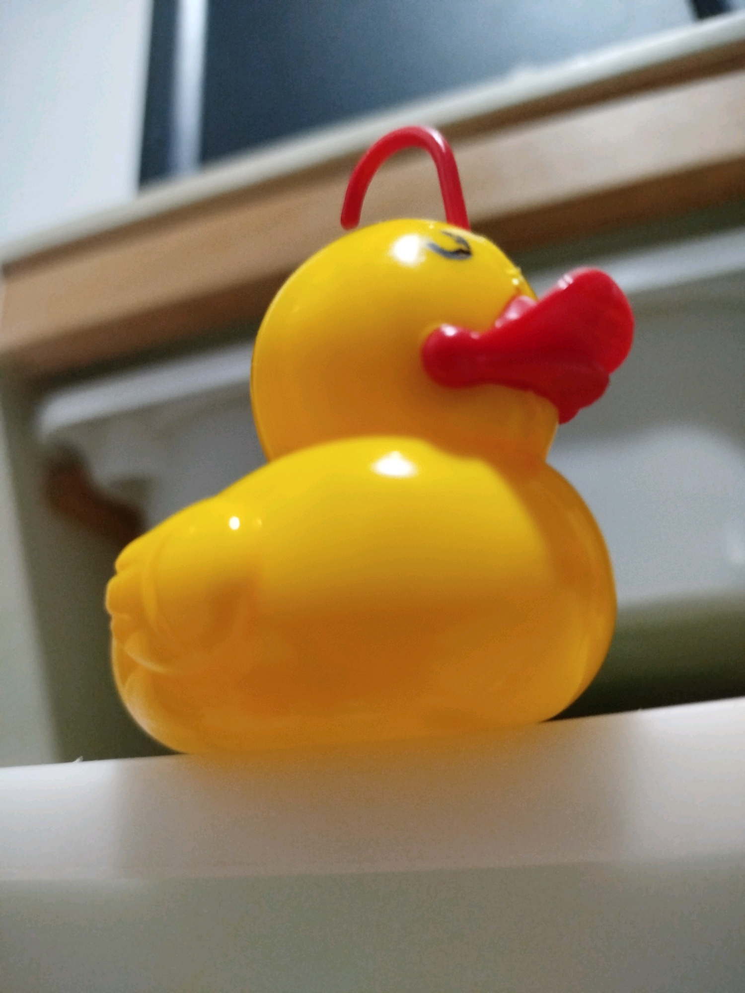 A yellow rubber duck toy with a red beak and a red handle on its head is positioned on a white surface. The duck is viewed from the side, with its body taking up most of the frame. The background is blurred, showing a wooden surface and what appears to be a wall with a dark rectangular shape. The lighting suggests an indoor setting. The image does not provide any specific details or context that would allow for its location in Mojoagung, Indonesia.