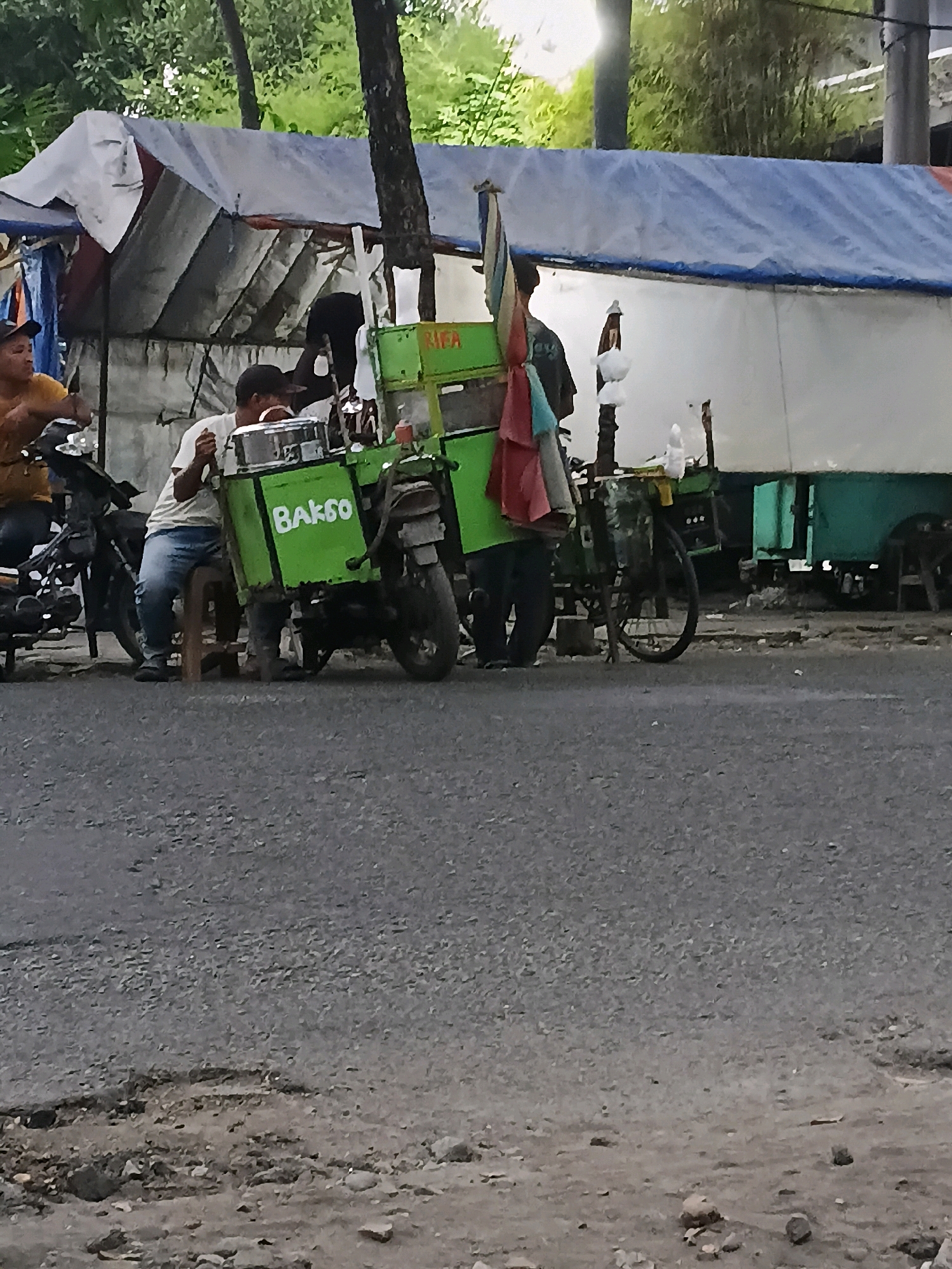 A street scene in Purwodadi, Indonesia, features a portable food stall, identified by the prominent green color and "Bakso" text on its side, which is mounted on a motorcycle. Two individuals are visible near the stall. One person, wearing a yellow shirt and a dark cap, is partially seen on the left, seemingly engaged with a motorcycle. Another person, dressed in a white t-shirt and blue jeans, is seated on a small stool, interacting with a metal container on the food stall. A third person, wearing dark clothing, is standing behind the stall. The stall itself is covered by a blue and white tarp, and additional tarps are draped over a structure behind it. The ground is paved with asphalt, showing signs of wear and tear, with some debris and rocks scattered along the roadside. Trees and foliage are visible in the background, suggesting an outdoor setting.