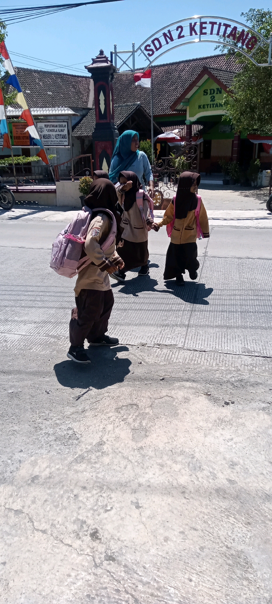 In Juwiring, Indonesia, a group of young students in uniform, accompanied by an adult, walk on a paved street. The students, three visible and one partially obscured, are wearing brown uniforms with matching headscarves. Two of the students, in the foreground and middle ground, carry light pink backpacks. The student in the foreground walks to the left, while the other two appear to be walking together to the right. The adult, wearing a light blue headscarf and shirt, walks behind the children, appearing to guide or supervise them. Behind the group, a school building with a red and white tiled roof and signage reading "SD N 2 KETITANG" is visible. The environment appears to be an outdoor, sunny day, with clear blue skies and some greenery visible in the background. The street is made of concrete, with visible lines and some cracks. Flags, including the Indonesian flag, are displayed near the school entrance.