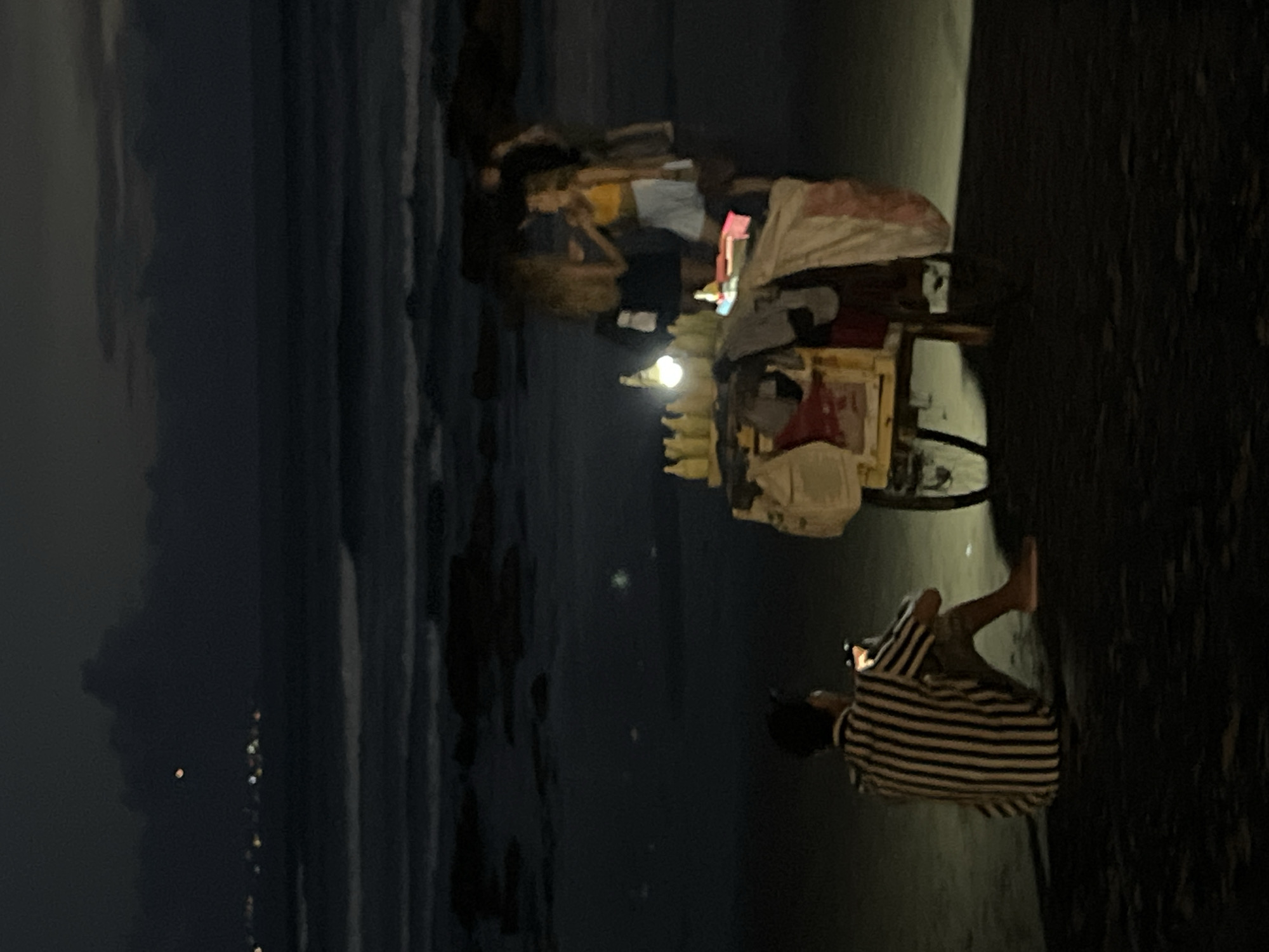 The image captures a nighttime scene on a beach in Canggu, Indonesia. In the foreground, a person wearing a striped shirt sits on the sand, looking at their phone. Nearby, there is a food cart lit by a lamp, with several people gathered around it. The background features the ocean and a dark, cloudy sky. The city lights are visible in the distance.