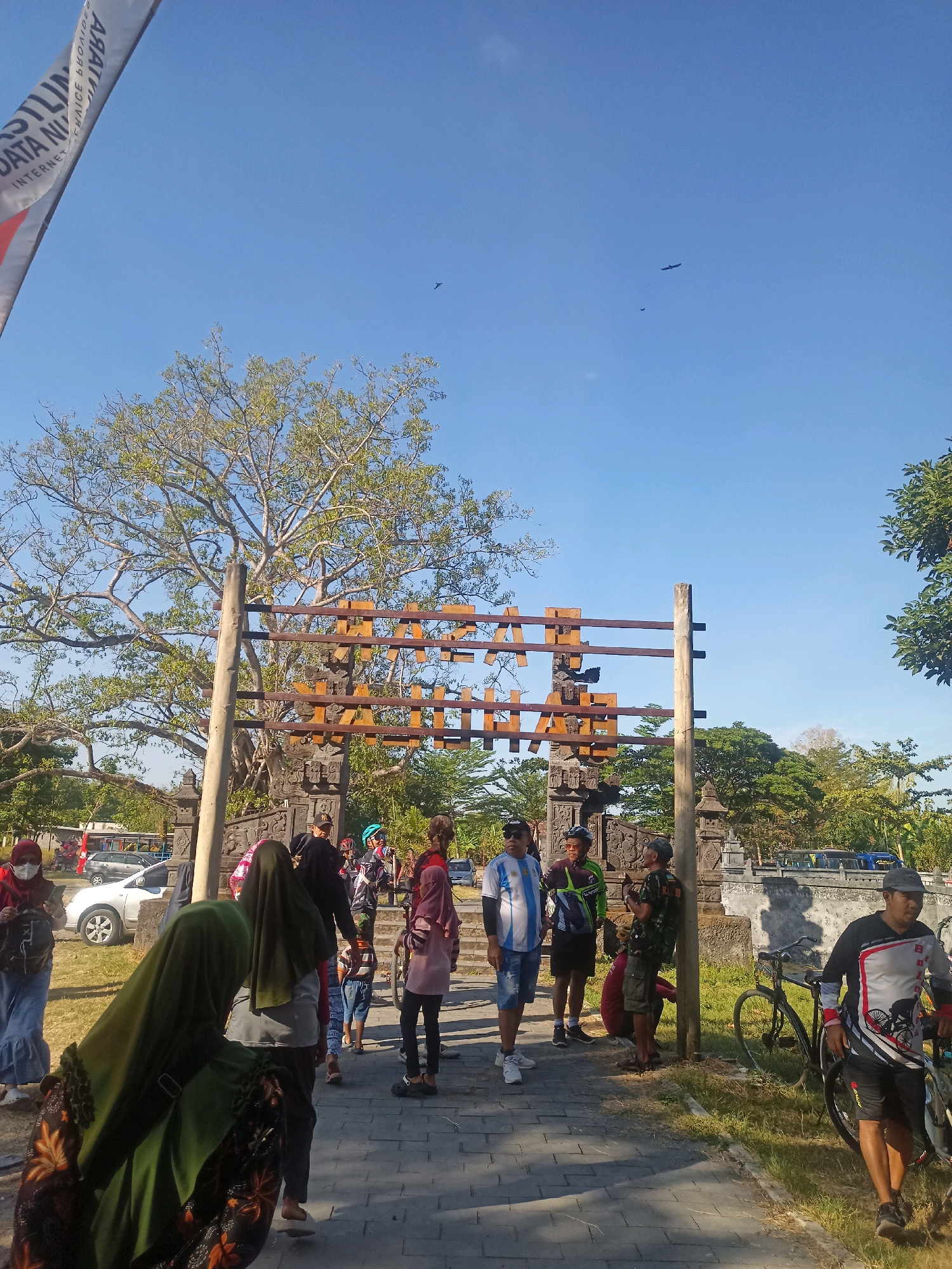 The image depicts a group of people gathered in front of an entrance archway in Karungan, Indonesia. The archway is constructed of wood and stone, with carved stone pillars supporting wooden beams that spell out "KARUNGAN" in large letters. A tall, leafy tree stands behind the archway.
Several individuals, some wearing cycling gear and helmets, are visible in the foreground and midground, suggesting a recreational outing, possibly a bike tour. One person is pushing a bicycle, and another is standing next to a parked bicycle. A woman in a green hijab and patterned dress is positioned prominently in the lower left of the frame, facing away from the camera. Other people are also seen in groups, some appearing to converse.
The environment is sunny with a clear blue sky. The ground is paved with interlocking stones. In the background, cars are parked and trees line the perimeter of the area. A flag is visible in the upper left corner. Two birds are flying in the sky. The overall impression is a bright, outdoor scene at a landmark or entrance.