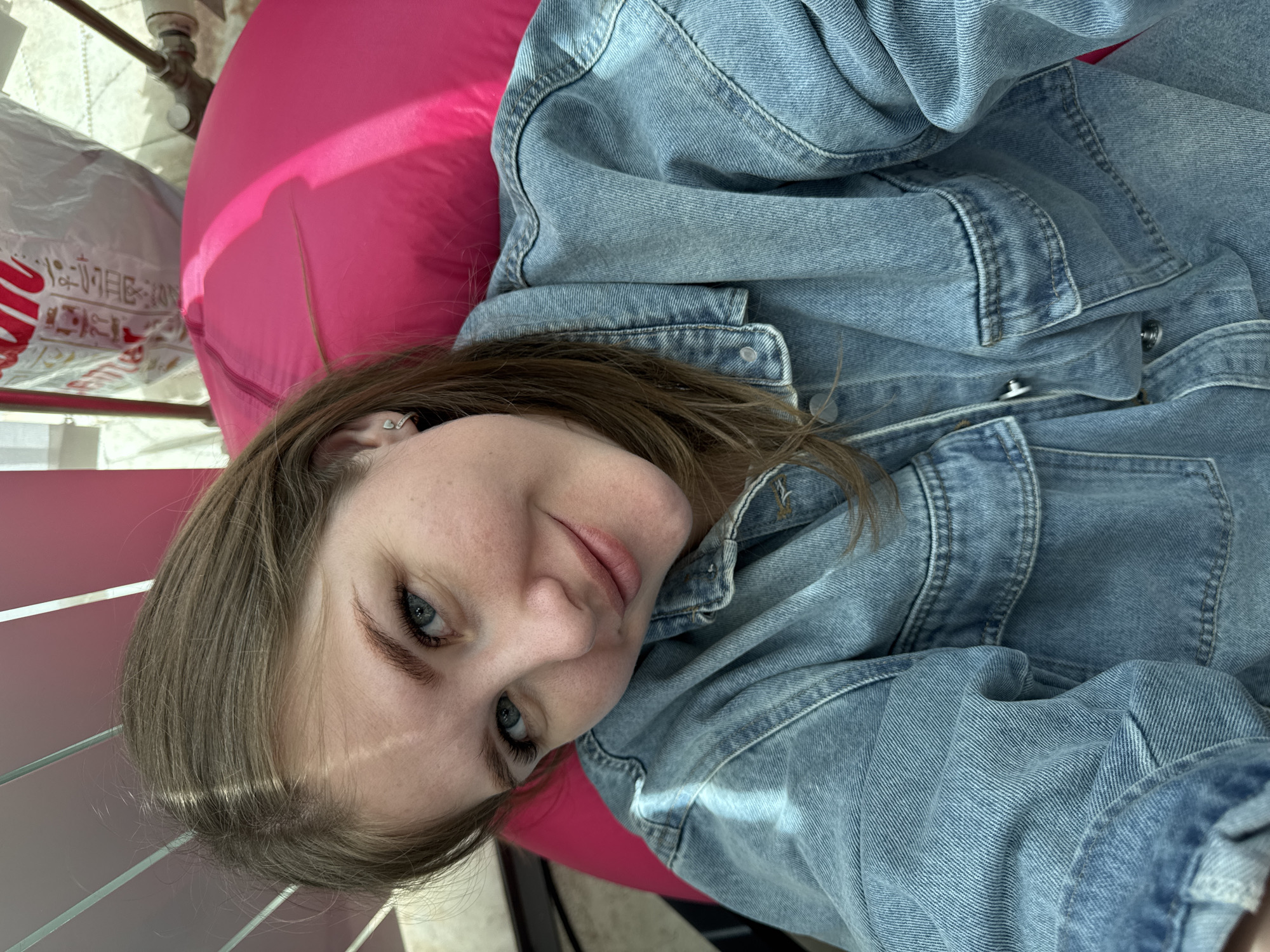 A young woman with light brown hair and blue eyes is lying on a bright pink beanbag chair, wearing a blue denim jacket. She is looking directly at the camera with a slight smile. A white plastic bag with red lettering is visible in the upper left corner of the image. The image was taken indoors, potentially near a window, given the light source. The location is Perm, Russia.