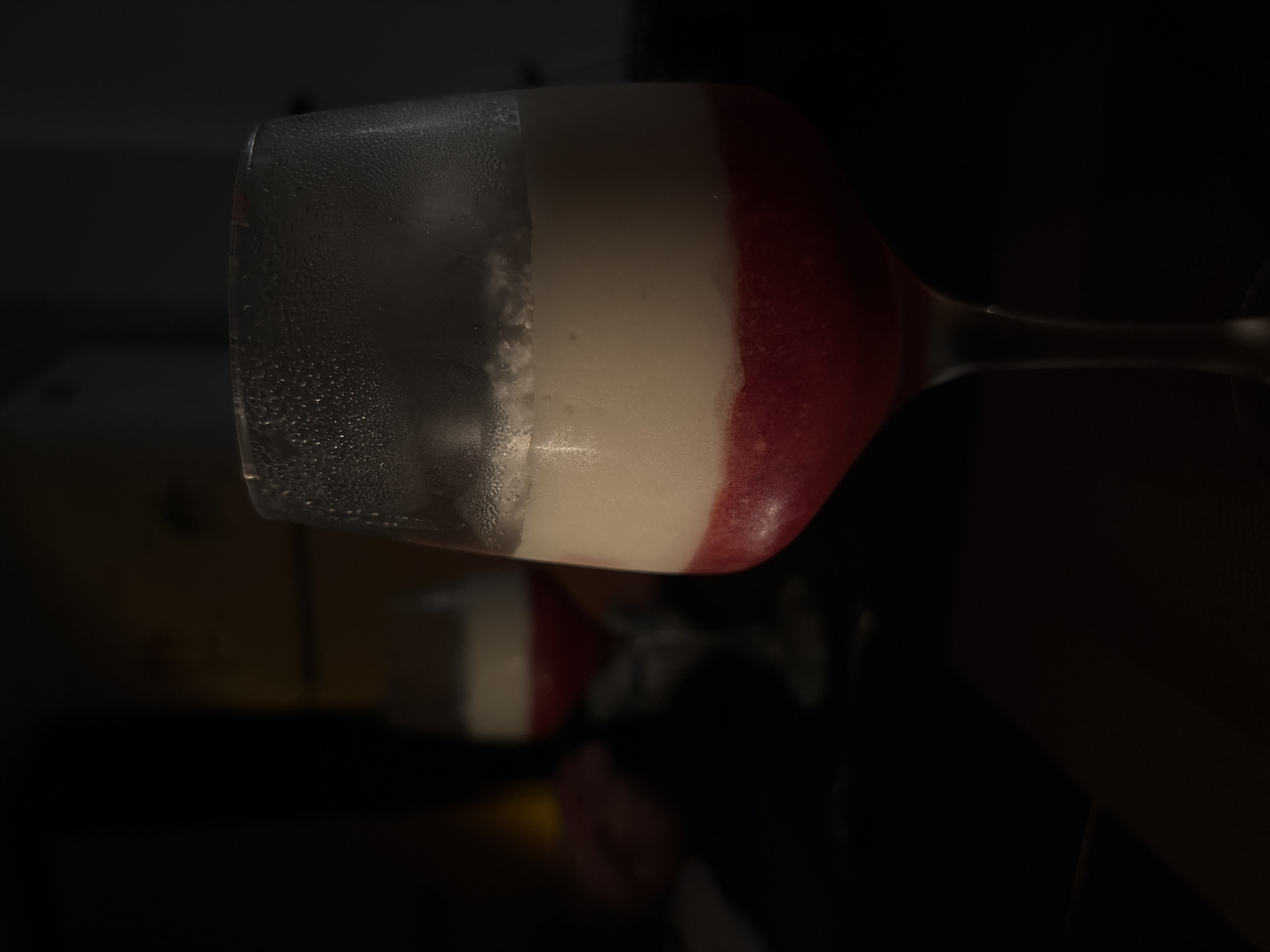 The image shows a close-up of a wine glass containing a layered dessert. The bottom layer is a vibrant red, appearing to be a fruit puree or jelly. Above this is a creamy white layer, possibly mousse or panna cotta. The glass itself is slightly frosted with condensation. In the background, another identical wine glass with the same layered dessert is visible, slightly out of focus, suggesting a restaurant or bar setting. The lighting is dim, with the focus on the foreground glass. The scene appears to be indoors, with dark, indistinct surroundings. There is no identifiable location in Bečići, Montenegro indicated in the image.