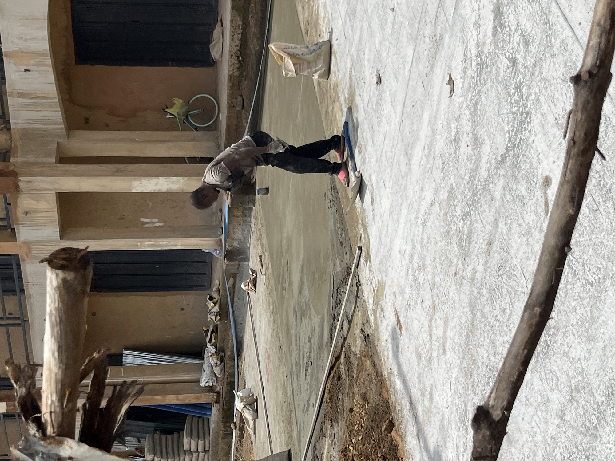 The image depicts a construction site in Bukuru, Nigeria. In the foreground, a person wearing a gray t-shirt, dark pants, and pink sandals is actively smoothing wet concrete with a trowel. The person is leaning over the concrete surface, focused on the task. Tools, including a trowel, are visible on the ground near the person. Several bags of cement are also present, with one large bag positioned to the right of the concrete area. The environment shows areas of exposed earth and a newly laid concrete surface. In the background, there is a building with an open doorway and a bicycle. The scene suggests ongoing construction work.