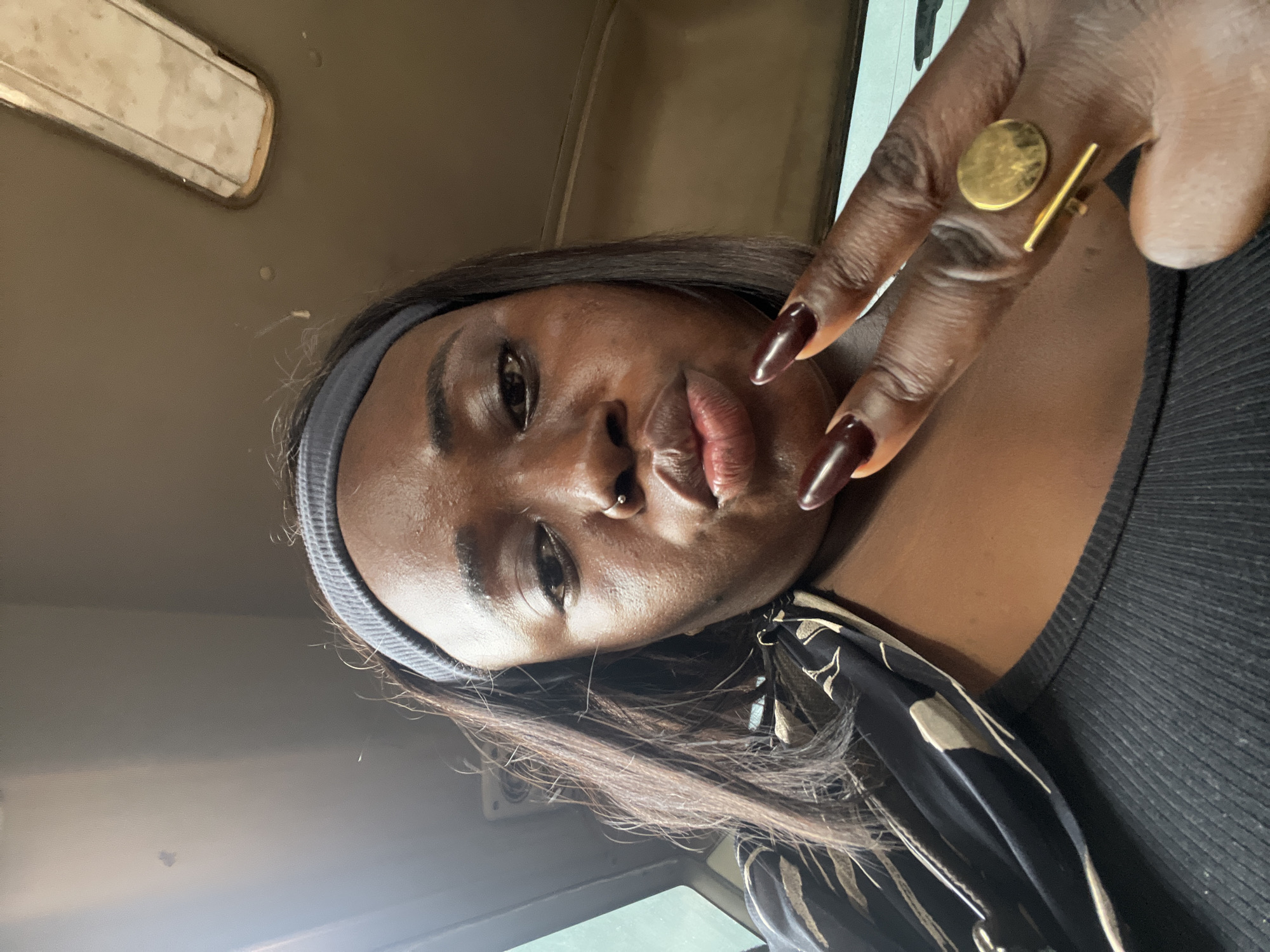 A close-up selfie of a Black woman with dark skin and long, straight dark brown hair is captured. She is wearing a dark gray headband and a black top with a ribbed texture. A black and tan patterned scarf is draped around her neck and shoulders. Her lips are pursed, and she has a nose ring. Her right hand is raised, displaying long, dark red-brown nails, and she is wearing a gold ring on her right index finger. The ring features a circular element and a horizontal bar. She appears to be in the interior of a vehicle, with a glimpse of a window and a ceiling light fixture visible in the background. The lighting suggests it is daytime. The environment is not detailed enough to definitively place it in Kugbo, Nigeria.