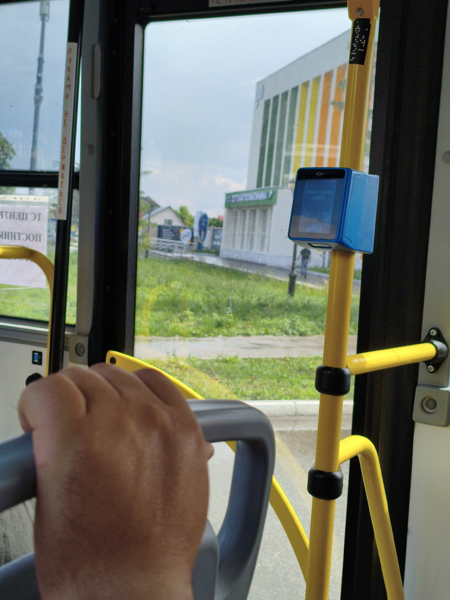 The image, taken from inside a bus in Orenburg, Russia, shows a person's hand gripping a gray handrail. In the foreground, a yellow metal pole with black rubber grips is visible, supporting a blue electronic fare payment device mounted at the top. The device has a screen displaying text in a language likely to be Russian. Through the bus window, a street scene is visible, including a building with colorful vertical stripes, a grassy area, and trees. The sky is overcast. The handrail and pole are typical of public transportation interiors designed for passenger safety.