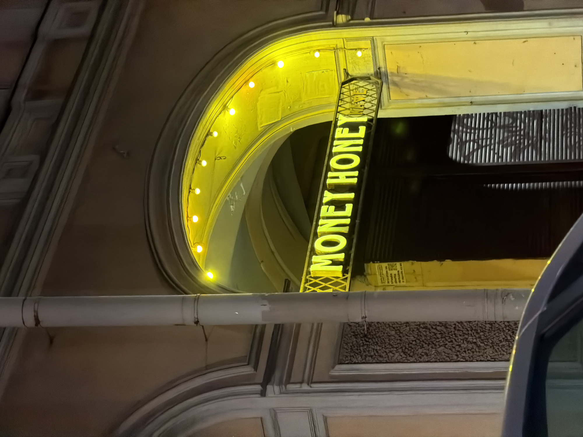 The image captures a night scene in Saint Petersburg, Russia, featuring a business with the illuminated sign "MONEY HONEY". The sign is prominently displayed, positioned between a white pipe running horizontally across the frame and an arched architectural feature adorned with yellow lights. The architecture is ornate and classic, suggesting an older building. Part of a vehicle is visible in the lower right of the frame.