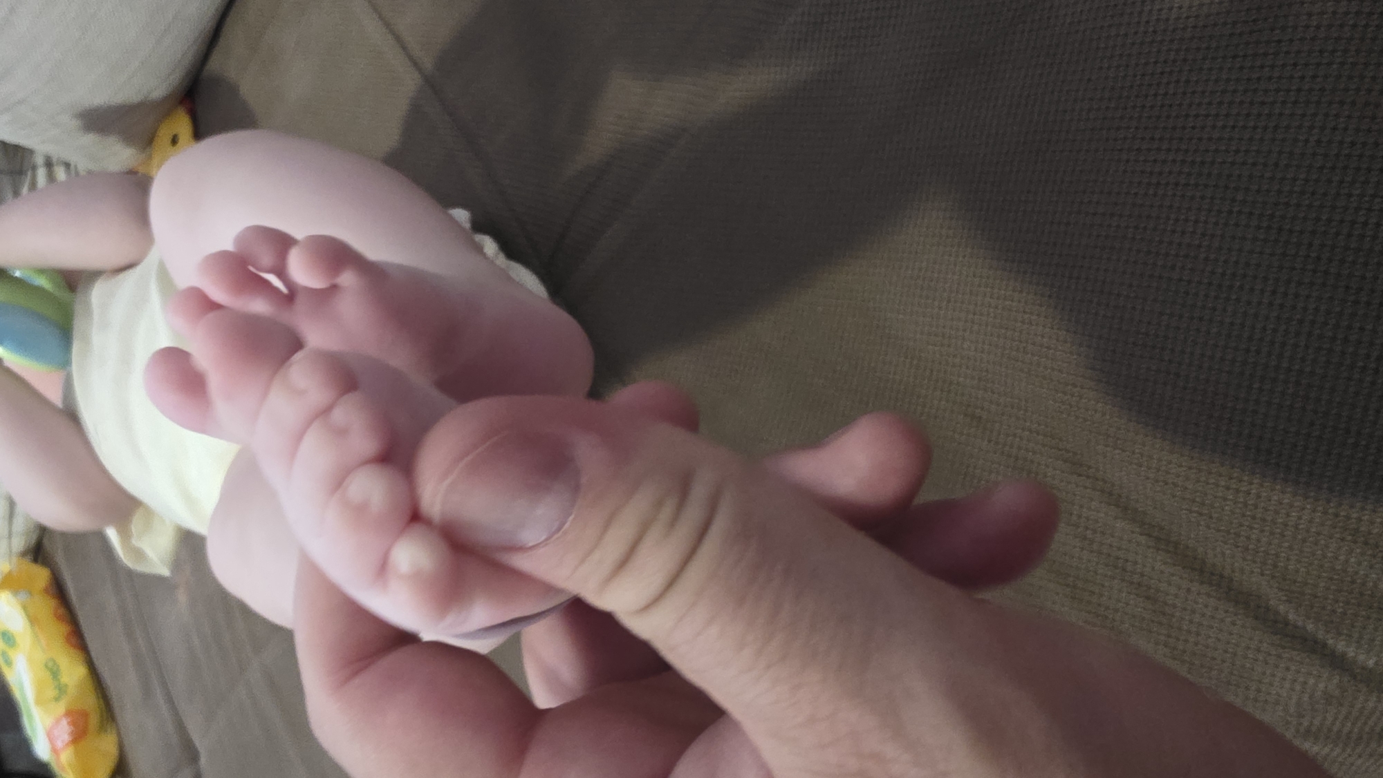 A person's hand is holding a baby's foot. The baby is lying on a brown couch and is wearing a diaper. A yellow object is visible in the background. The location cannot be determined from the image.