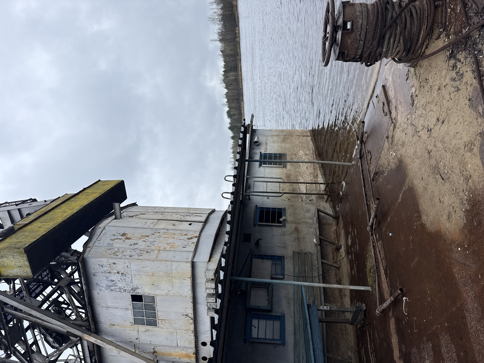 The image depicts a decaying industrial scene. A large, weathered metal structure with peeling paint and multiple windows sits adjacent to a body of water, possibly a river or lake, with trees visible on the opposite shore. The metal structure appears to be part of a larger complex, with a crane-like arm and steel lattice framing on one side. A heavy, rusted metal wheel and coiled cable are prominent in the foreground, suggesting the structure's former use for heavy lifting or transport. The overall atmosphere is one of abandonment and decay. This location appears to be within the Chornobyl Exclusion Zone in Ukraine, where industrial facilities have been left abandoned due to the 1986 nuclear disaster. The building has four windows on one side with blue frames.