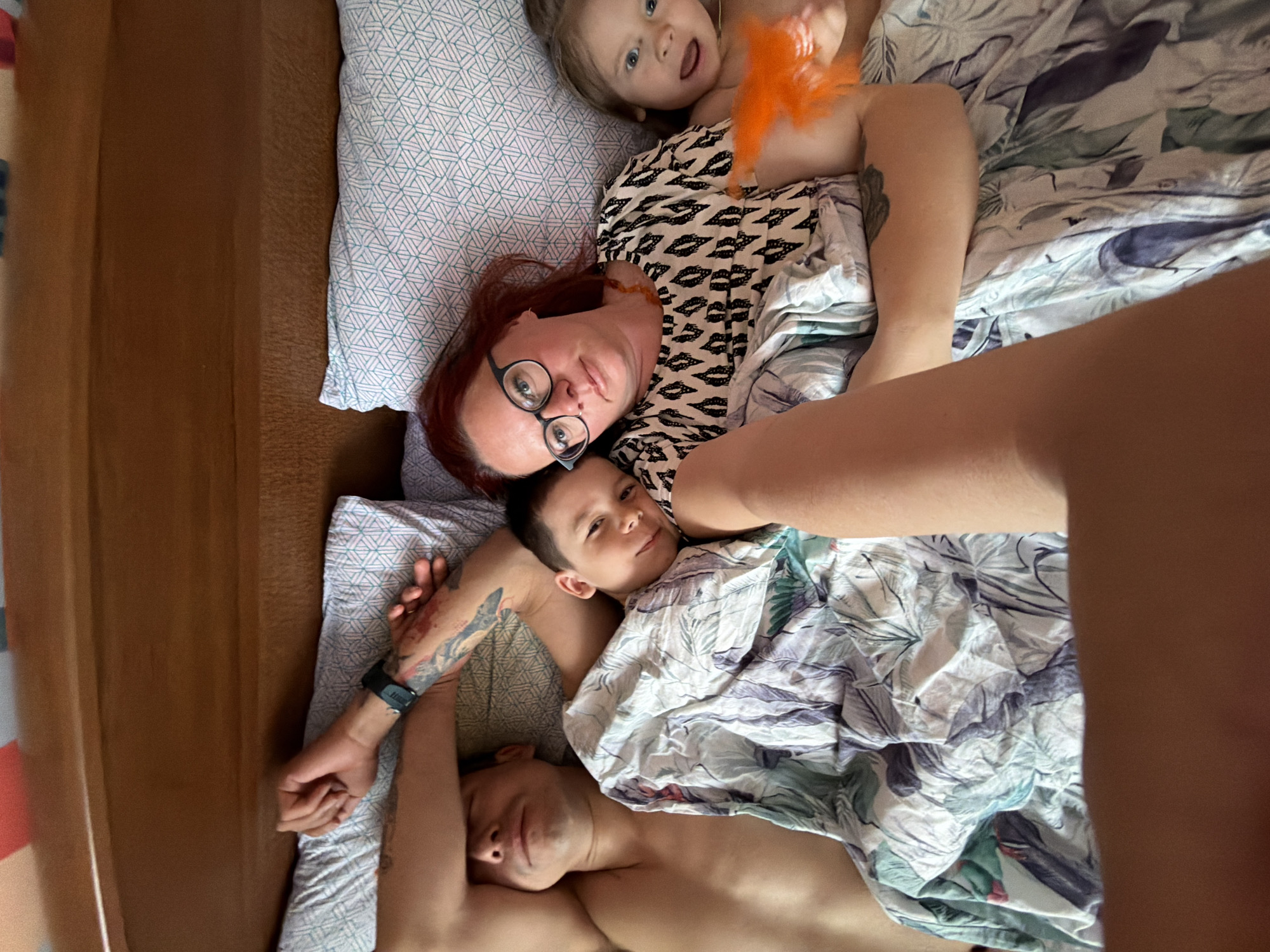 A high-angle shot captures a family of four, including two adults and two children, relaxing together in a bed in Chaikovsky, Russia. The perspective is from above, looking down on the subjects, who are arranged horizontally across the frame.
In the upper central part of the image, a woman with reddish-brown hair and glasses lies on a pillow with a geometric light blue and white pattern. She is wearing a black and white patterned top and looks directly at the camera with a soft expression. Beside her, to the upper left, a young child, a girl with light brown hair, is also looking up at the camera with a wide-eyed, slightly open-mouthed expression, holding an orange feathery toy. Her arm is visible, showing bare skin.
Below the woman, to the left, an older child, a boy, rests his head on the woman's arm, looking towards the camera with a gentle smile. He is partially covered by a duvet with a green and blue botanical pattern. Further down, an adult male lies at the bottom of the frame, seemingly asleep or resting with his eyes closed. He has tattoos on his left arm, and a dark-colored watch is visible on his wrist. His chest is bare.
The bed appears comfortable, with soft pillows and patterned bedding. The general lighting suggests daytime, creating a warm and intimate atmosphere. The family appears to be enjoying a relaxed moment together. No visible text is present in the image.