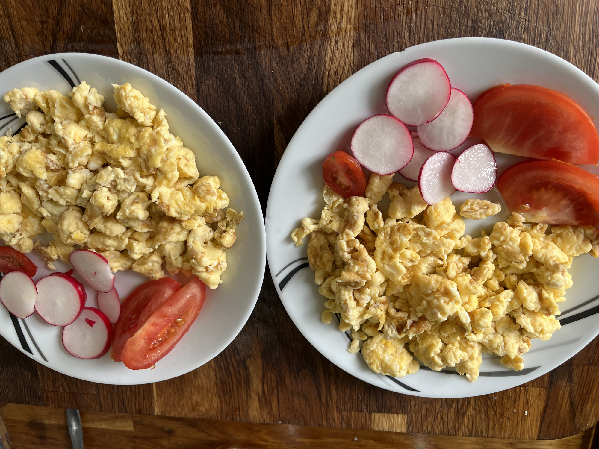 The image presents an overhead view of two white, round plates, each holding a serving of scrambled eggs accompanied by sliced vegetables. The plates are set on a textured wooden surface, possibly a cutting board or a rustic table, which serves as the background.
Both plates are filled with yellow, fluffy scrambled eggs, appearing soft and lightly cooked. On the left plate, several thin slices of radishes, characterized by their white interior and pinkish-red edges, are arranged along the bottom left side. Two wedges of red tomato are also placed next to the radishes.
Similarly, the plate on the right features scrambled eggs with a garnish of radishes and tomatoes. Multiple radish slices are distributed on the top left and bottom right sections of the plate, showcasing their vibrant colors. Alongside them are red tomato wedges and a smaller half of a cherry tomato. Both plates feature a subtle black and grey linear design along their rims, which is more clearly visible on the right plate.
The scene suggests an indoor setting, likely a kitchen or dining area, with the meal prepared for consumption, presumably breakfast or brunch. No people are visible, and no specific activity is taking place beyond the presentation of the food. There is no visible text in the image. The overall impression is one of a simple and wholesome meal.