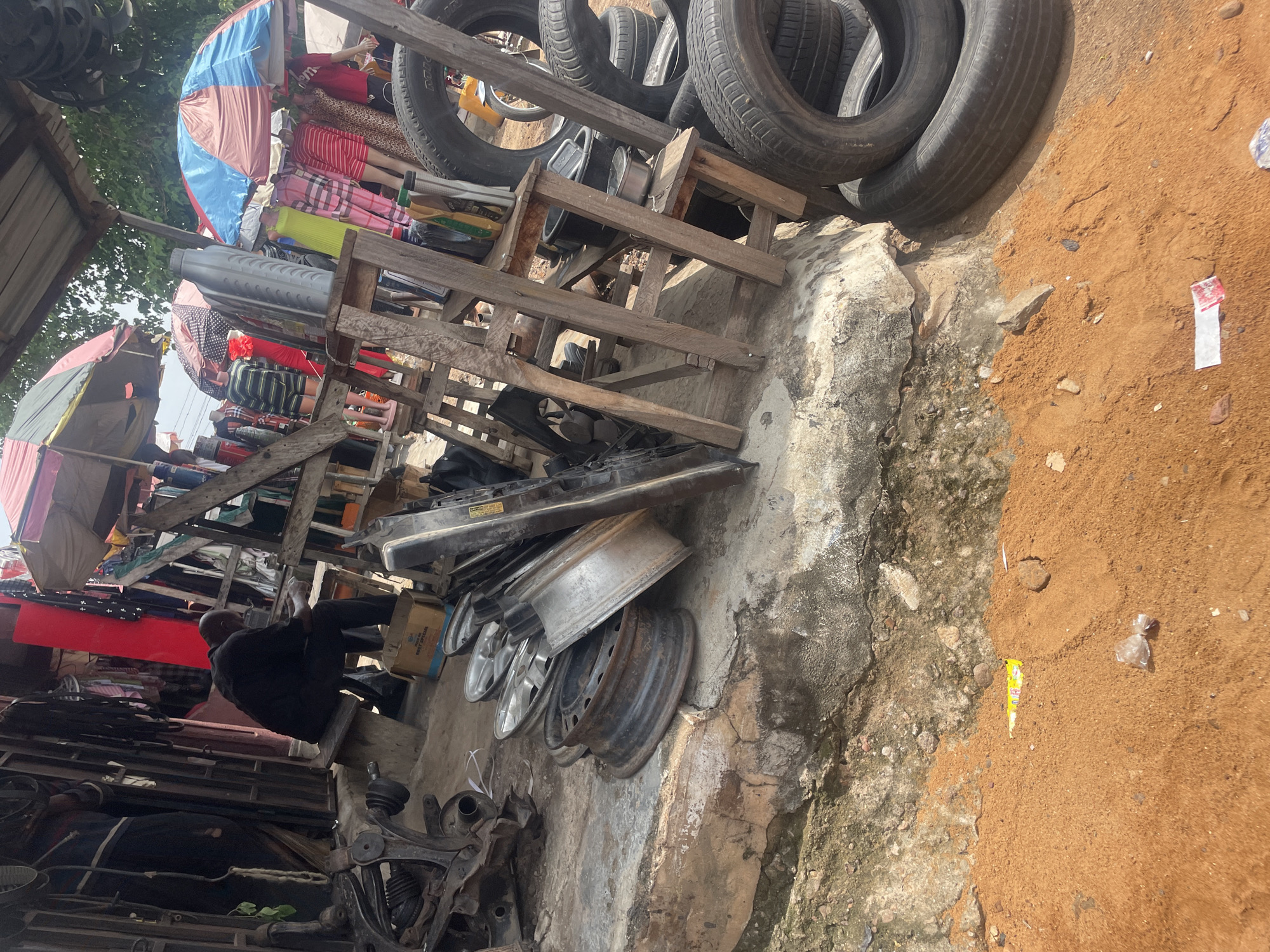 This image captures a street scene in Lokoja, Nigeria, likely a marketplace or a repair shop. In the foreground, a pile of car tires, some stacked, others individually placed, dominates the right side. Among them, a section of a concrete structure with rough, textured surfaces is visible. To the left, various car parts, including what appears to be suspension components and exhaust systems, are scattered on the ground. Further back, a man wearing dark clothing is seated on a low stool, facing away from the camera, seemingly occupied with something in his hands. Behind him, shelves are laden with items, and fabric, possibly clothing, is displayed. A red metal structure, possibly part of a stall or building, stands prominently in the background. Multiple umbrellas, some colorful, are set up, suggesting shelter from the sun. The ground is a mix of concrete, dirt, and sand, with some scattered debris. The overall impression is one of a busy commercial area with a focus on automotive parts and possibly clothing sales.