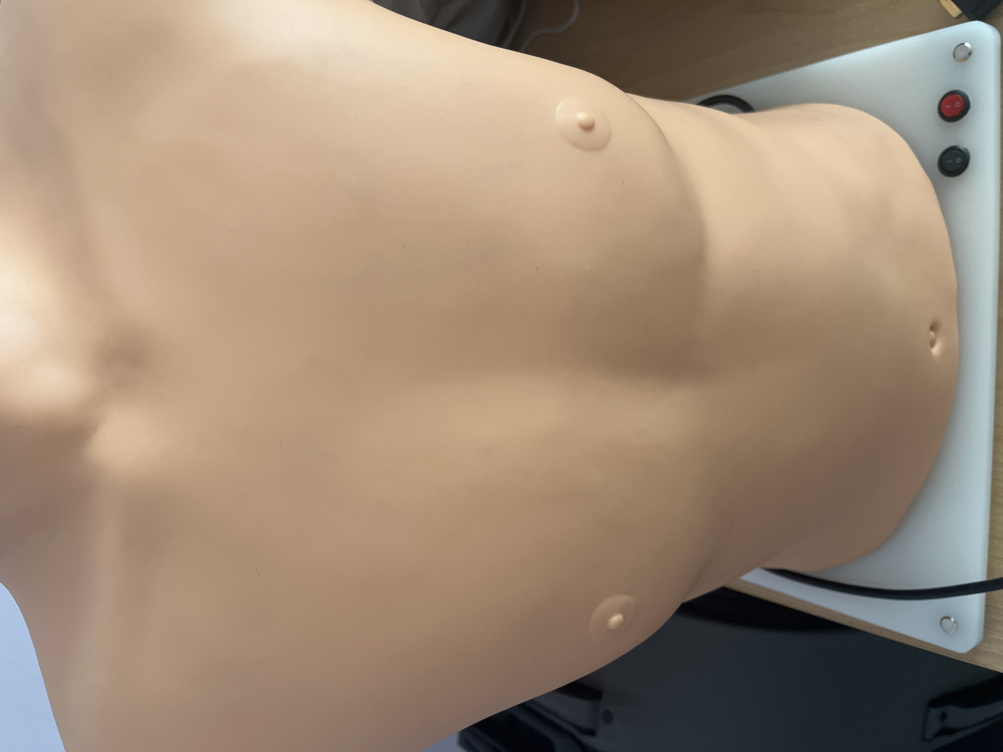 The image displays a medical training mannequin's torso, positioned horizontally. The mannequin is skin-toned, depicting a human-like upper body with features resembling a female chest, including two prominent nipples and a distinct belly button (navel). Its surface appears smooth.
The mannequin rests on a white, rectangular base. On the right side of this base, there are several circular controls: a red switch or button and a black switch or button are clearly visible, along with a silver metallic stud or screw. Black electrical wires or cables are connected to the base, indicating it is an electronic training device.
The setup is placed on a light-colored wooden surface, likely a desk or table, with a dark, indistinct object partially visible in the background beneath the base. The lighting is even, suggesting an indoor environment such as a medical training facility or classroom. No text is visible in the image.