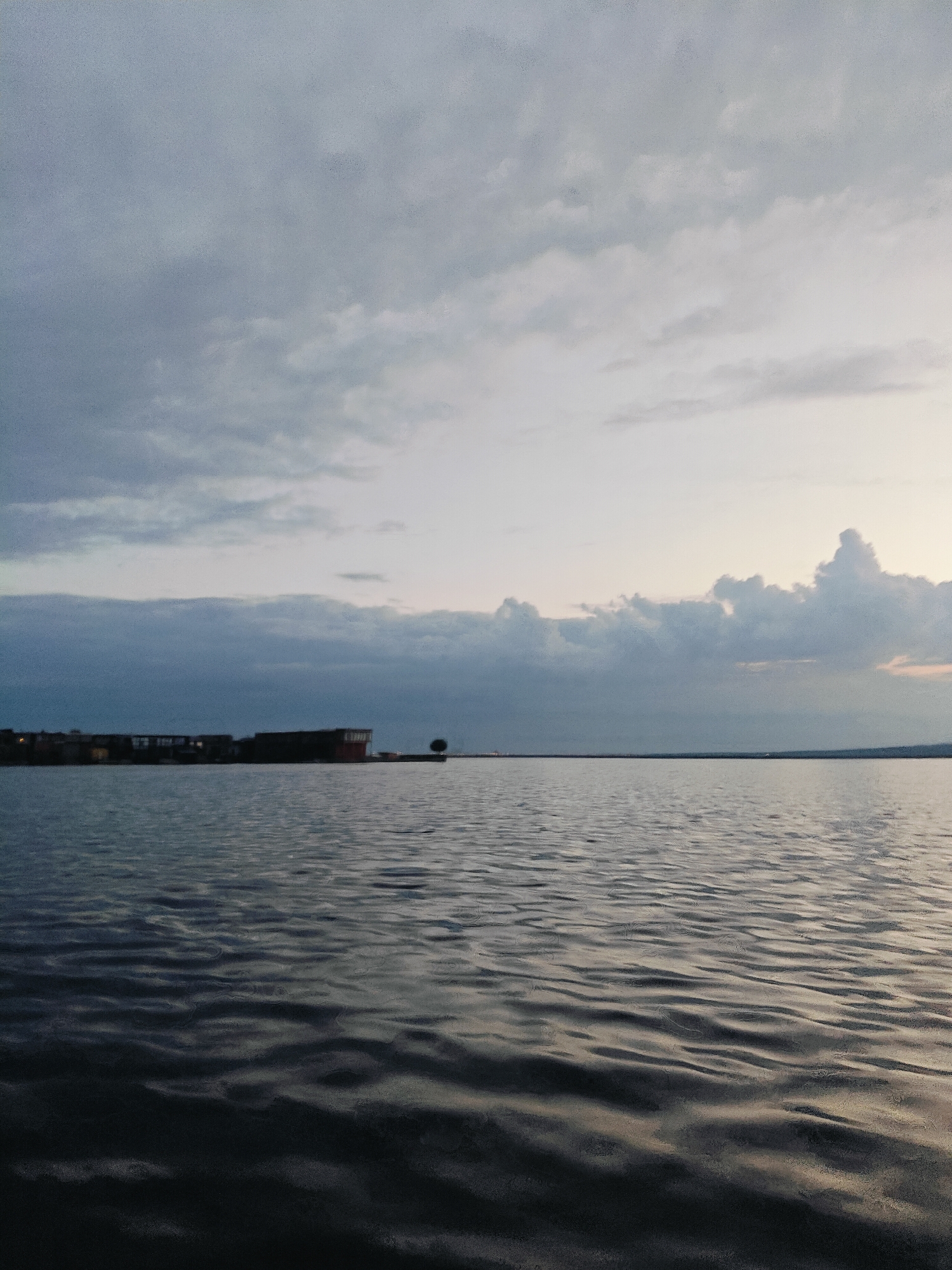 The media file depicts a serene, wide-angle view of a body of water, likely a river or lake, with a subdued cloudy sky overhead. The water's surface is disturbed by gentle ripples, reflecting the muted light. In the distance, along the horizon, a line of industrial or residential structures is visible, appearing somewhat elongated and dark against the water. To the right of these structures, a solitary tree stands on what appears to be a dock or a small island. The overall mood is calm and slightly melancholic, possibly indicative of dusk or an overcast day.
The lighting suggests it is late afternoon or early evening, with the sky transitioning between shades of gray, white, and a hint of pale orange or pink on the far right, possibly from a setting sun. The clouds are varied, with some appearing dense and dark, while others are lighter and more diffused. The location, identified as Chaikovsky, Russia, places this scene in a context of a potentially industrial or port area, given the structures on the shoreline. There is no activity or event clearly taking place; the image captures a moment of stillness. No visible text is present in the media file.