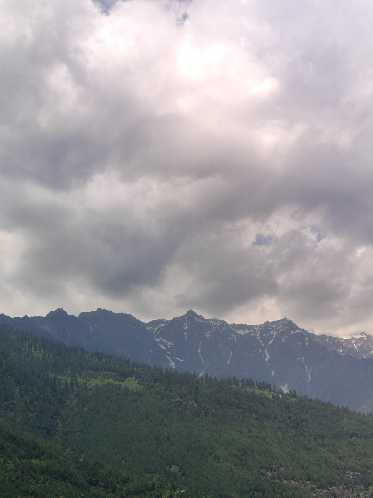 The image shows a vast mountain range under a cloudy sky. The lower slopes of the mountains are covered in dense green pine trees, with patches of lighter green indicating clearings or different vegetation. Higher up, the mountains become rocky and jagged, with streaks of snow visible on their peaks and sides. The sky is filled with dramatic, grey and white clouds, with some areas of brighter light suggesting the sun is partially breaking through. The overall scene is one of natural grandeur and a slightly overcast, possibly cool, weather condition. The location is Vashisht, India.