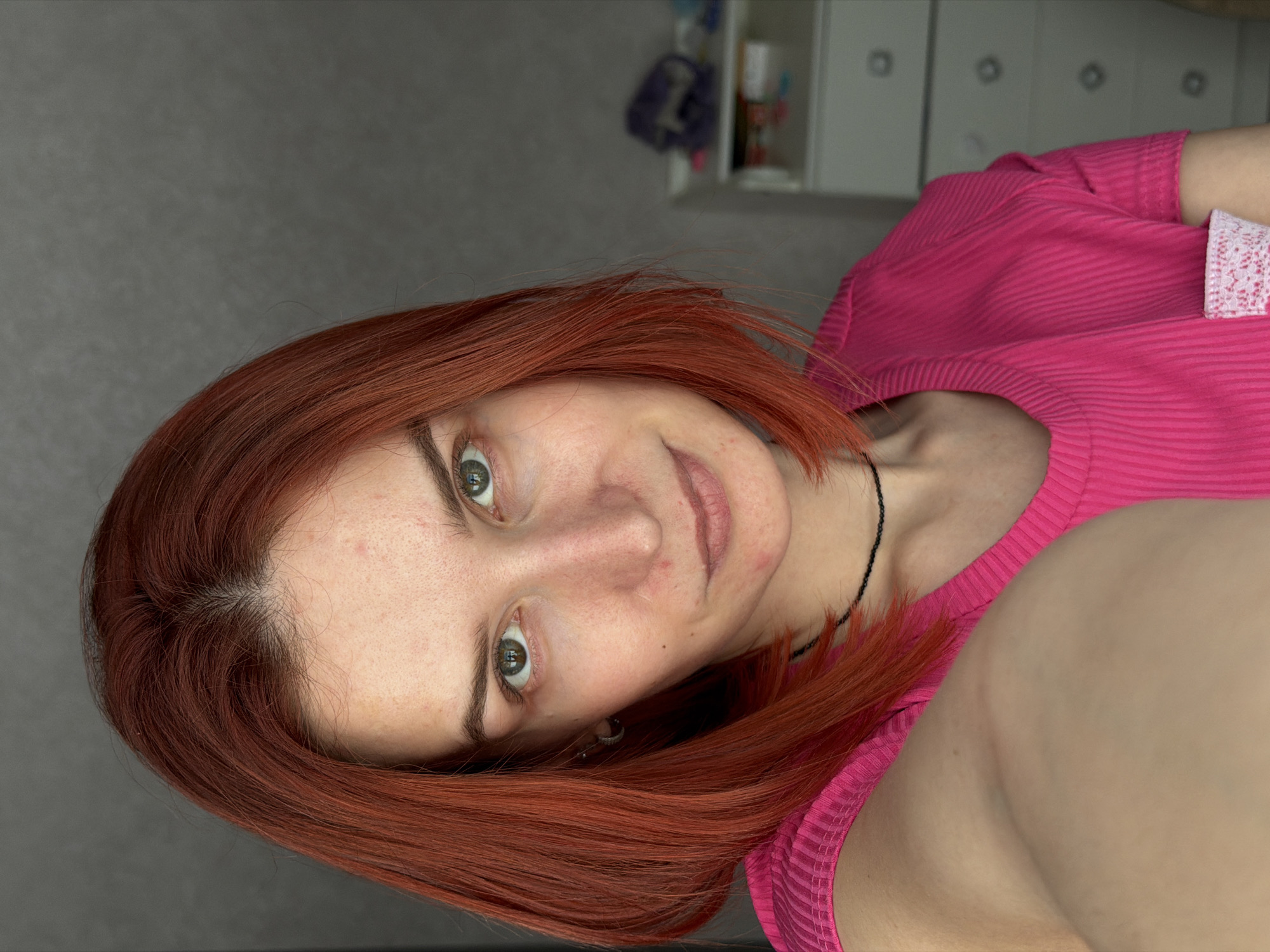 A woman with red hair and green eyes, wearing a pink ribbed shirt, is posing for a selfie. She is wearing a black choker necklace and has multiple piercings in her left ear. The background includes a gray wall and a white cabinet with silver knobs. The image appears to have been taken indoors. The location in the city of Chaikovsky, Russia is not evident from the visual information provided.