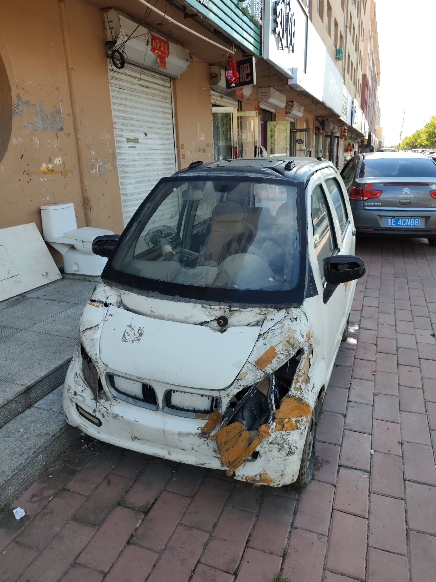 In Lindian, China, a damaged white electric car sits parked on a brick sidewalk next to a building with an orange facade and a roll-down metal shutter. The car's front end is heavily damaged, with the bumper and hood bent and torn, revealing orange padding underneath. The passenger side mirror is also broken. Behind the damaged car, a gray Citroën sedan is parked. The building appears to be a commercial establishment, with signs in Chinese visible above the storefront. To the left of the damaged car, a white toilet and a large white flat object are leaning against the building. The scene suggests an accident or disuse of the electric vehicle.