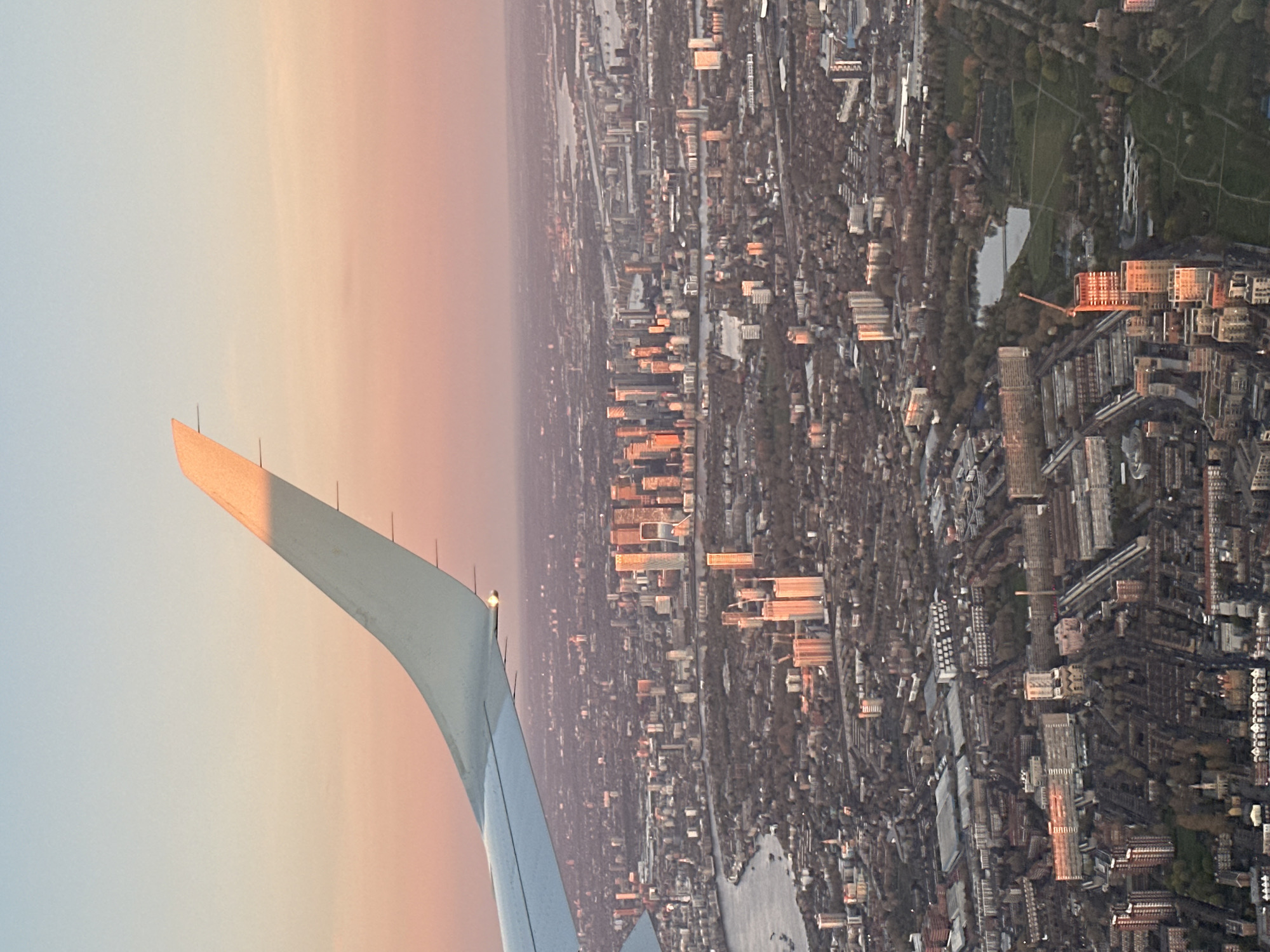 The image shows an aerial view from an airplane over London, United Kingdom. The wing of the plane is visible on the left side of the image. The cityscape is densely populated with buildings of various sizes, interspersed with green spaces. The buildings are reflecting the warm light of the setting or rising sun. A river winds through the city. Hyde Park is visible.
