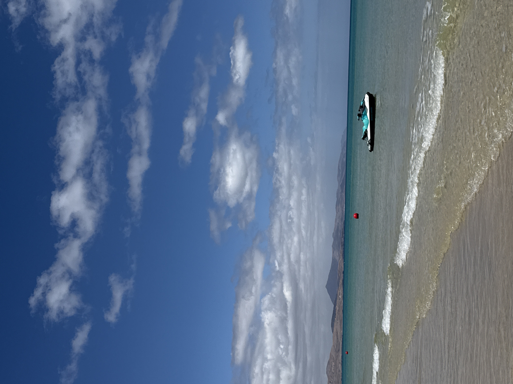This image captures a serene coastal scene, likely in Pájara, Spain, on a clear and sunny day. The dominant feature of the upper left portion of the frame is a bright blue sky dotted with scattered, elongated white clouds, suggesting a pleasant, breezy atmosphere.
The right side of the image displays a calm, turquoise ocean meeting a light-colored sandy beach. Gentle, frothy waves are seen breaking softly along the shore, creating a diagonal line across the lower right. A white and blue/teal jet ski is floating peacefully in the shallow water, positioned near the center of the water body. Two small red spherical buoys are visible in the water; one is closer to the jet ski, and another is further down the beach towards the foreground.
In the background, a range of mountains or hills rises, their arid, light brown slopes indicating a dry climate. The mountains appear somewhat hazy in the distance. The overall impression is one of tranquility and leisure, with no human activity directly depicted. The time of day appears to be mid-day given the bright lighting. No visible text is present in the image.