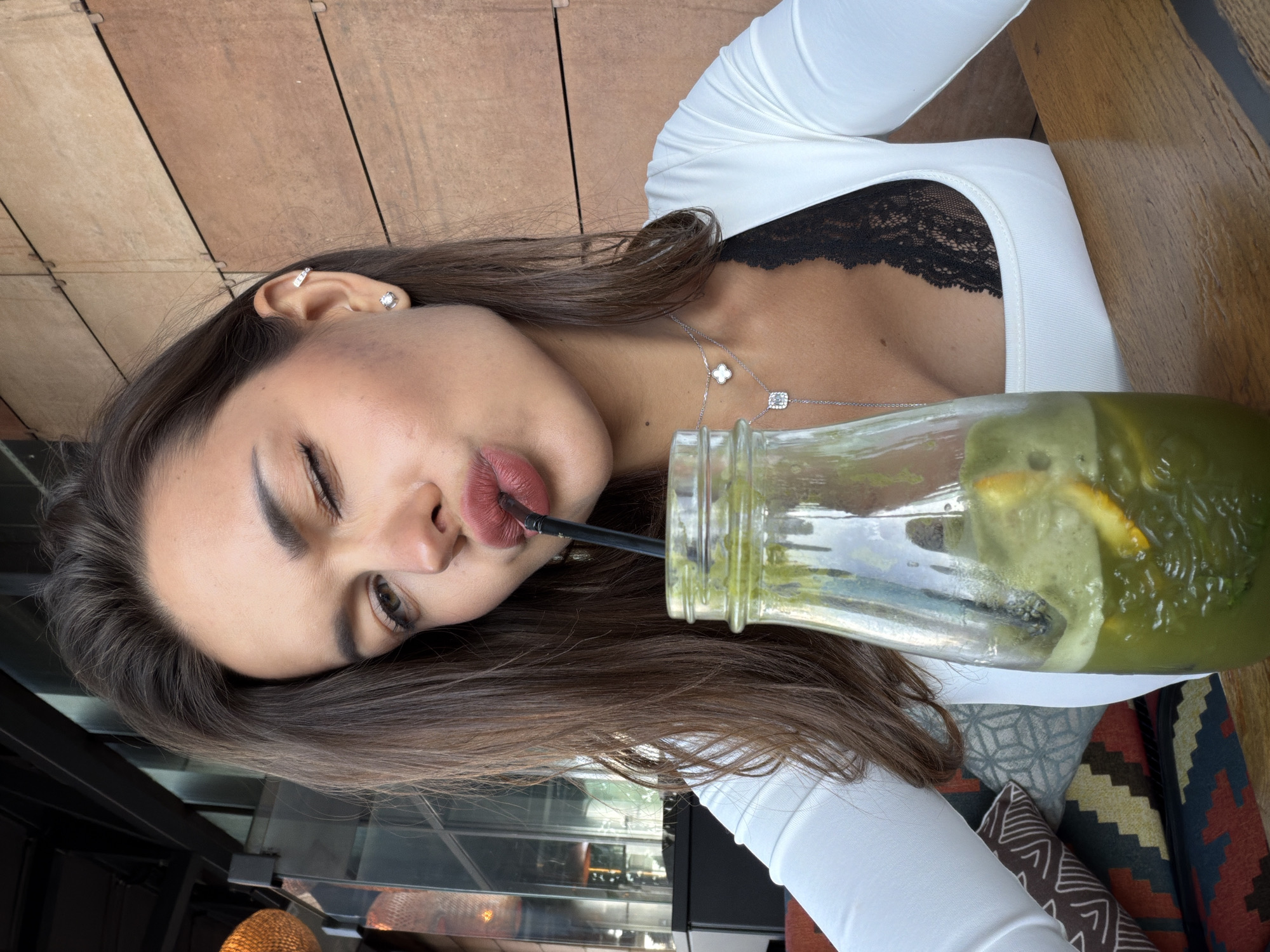 A young woman with dark hair is taking a selfie while drinking a green beverage from a glass bottle with a straw. She is wearing a white top with black lace detailing underneath. She is also wearing a necklace with a clover pendant. The background appears to be a restaurant or cafe with wooden walls and seating. The lighting suggests it is daytime. The image is taken from a slightly low angle, looking up at the woman. The scene is set in Saint Petersburg, Russia.