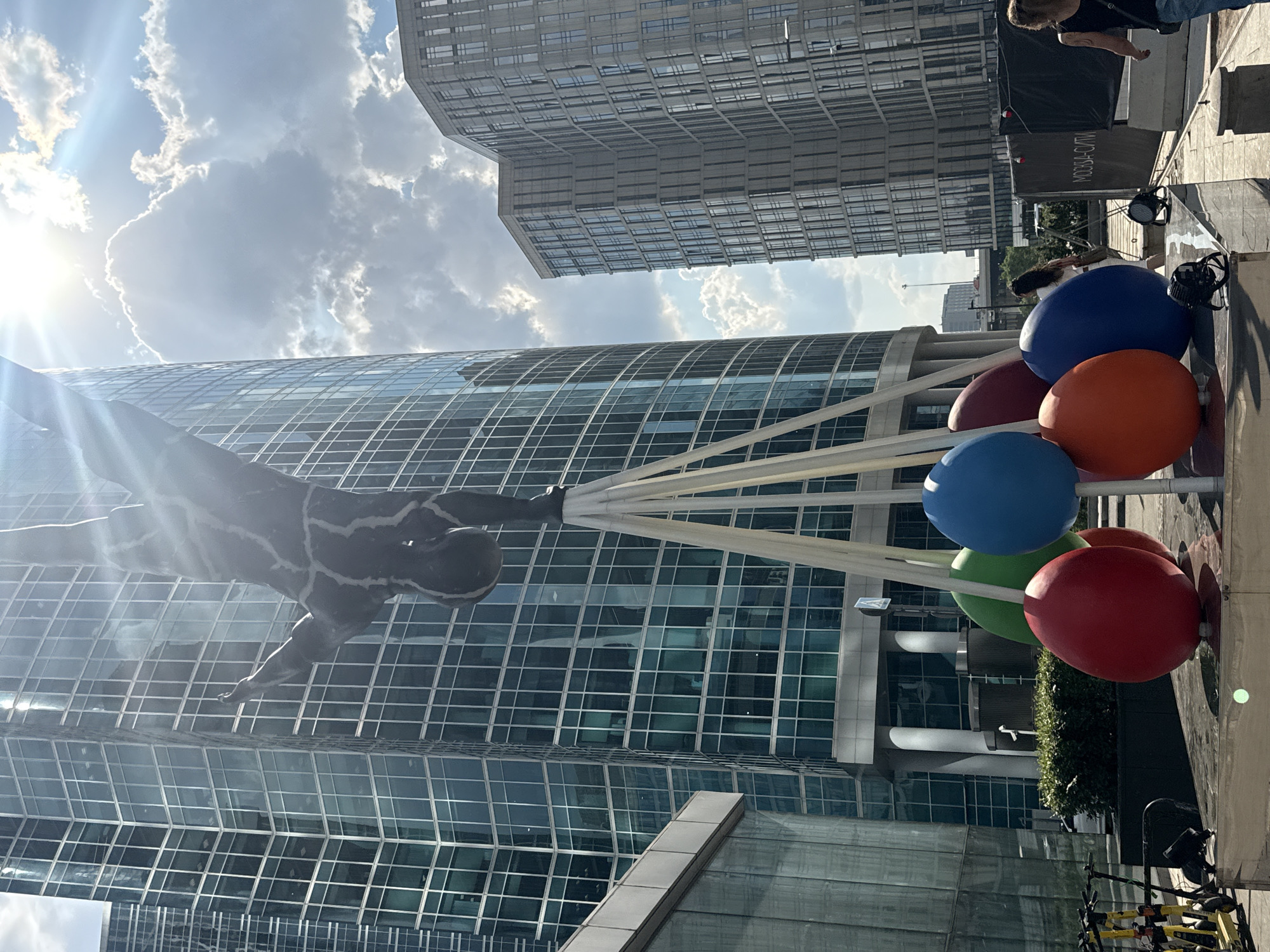 The image depicts a public art installation in Moscow, Russia. The sculpture, a dark, textured human figure performing a handstand, is suspended by a series of white poles. Below the sculpture, a cluster of large, brightly colored spherical objects—red, orange, blue, green, and maroon—are arranged on a platform. In the background, modern skyscrapers with reflective glass facades dominate the skyline, suggesting an urban, business district environment. The sun is visible in the upper right quadrant, partially obscured by clouds, casting a bright light and lens flare. Several electric scooters are parked on the left side of the frame. On the right, a banner with Russian text, "МОСКВА-СИТИ," is visible, confirming the location as Moscow City. Two individuals are present: one woman walking away from the camera towards the right, and another woman in the middle ground walking leftward. The overall scene is a juxtaposition of art, architecture, and urban life.