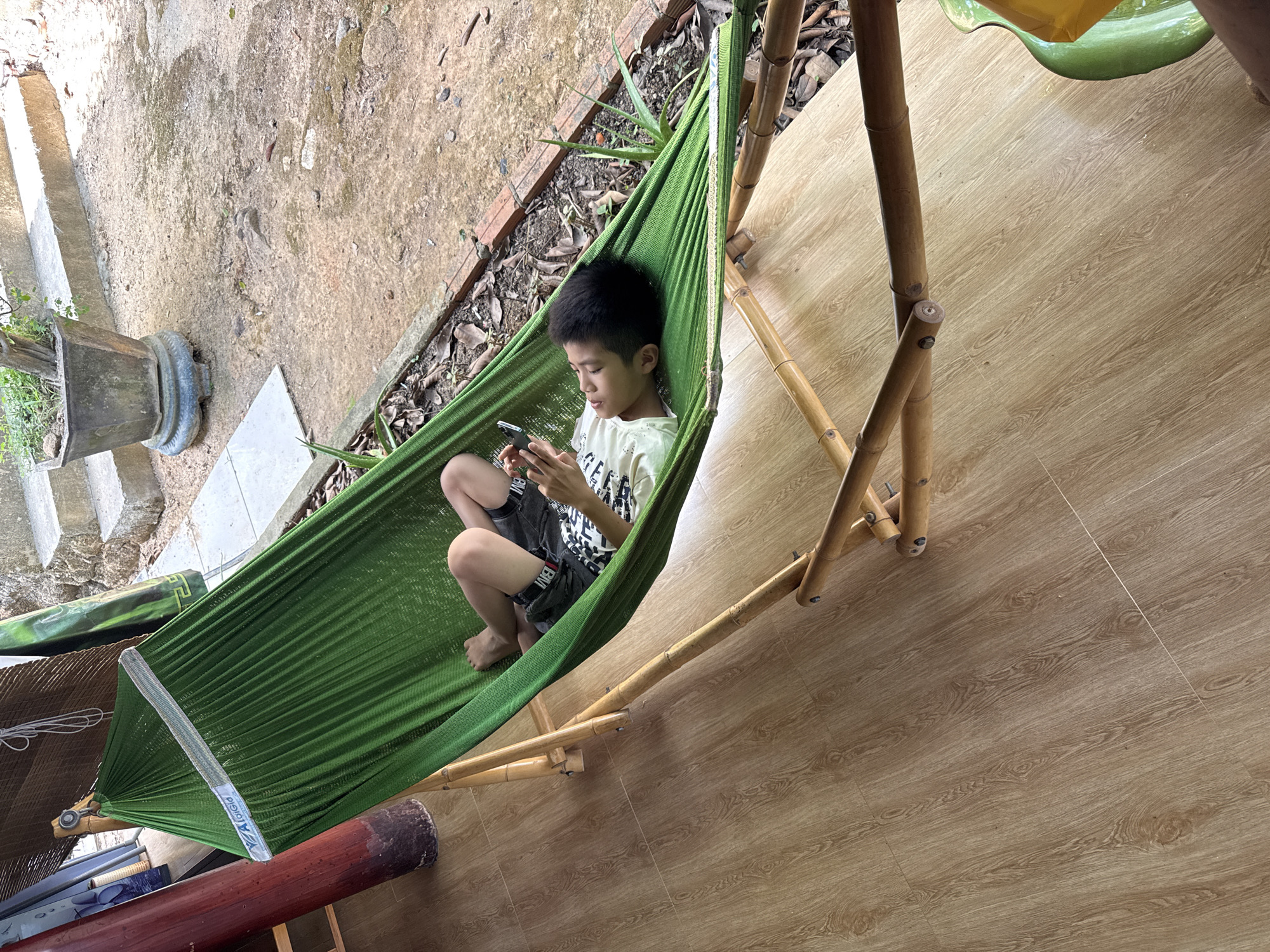 A young boy is captured from an overhead perspective, relaxing in a green fabric hammock outdoors in Tay Nha Trang Ward, Vietnam. He appears to be of Asian descent and is lying on his back, propped up slightly, intently focused on a black smartphone held in both hands. He is wearing a light-colored t-shirt with some illegible text and dark shorts that display the word "LEVI" on the leg.
The hammock itself is vibrant green and stretched between a natural bamboo stand on one side and an unseen anchor point on the other. A white label with blue text, including "LaLaKid", is visible on the hammock's spreader bar.
The setting is a transitional outdoor area. To the left of the hammock, there is a concrete ground or path, showing signs of wear and moisture, with a dark potted plant and some steps in the background. To the right, beneath the bamboo stand and extending further right, is a clean, light brown floor with a wood-grain pattern, possibly tiles or laminate, suggesting a patio or an adjacent indoor space. Scattered dried leaves and some small plants are visible near the concrete path. The scene is brightly lit by natural daylight, indicating a clear or partly cloudy day, and the boy appears calm and absorbed in his activity.