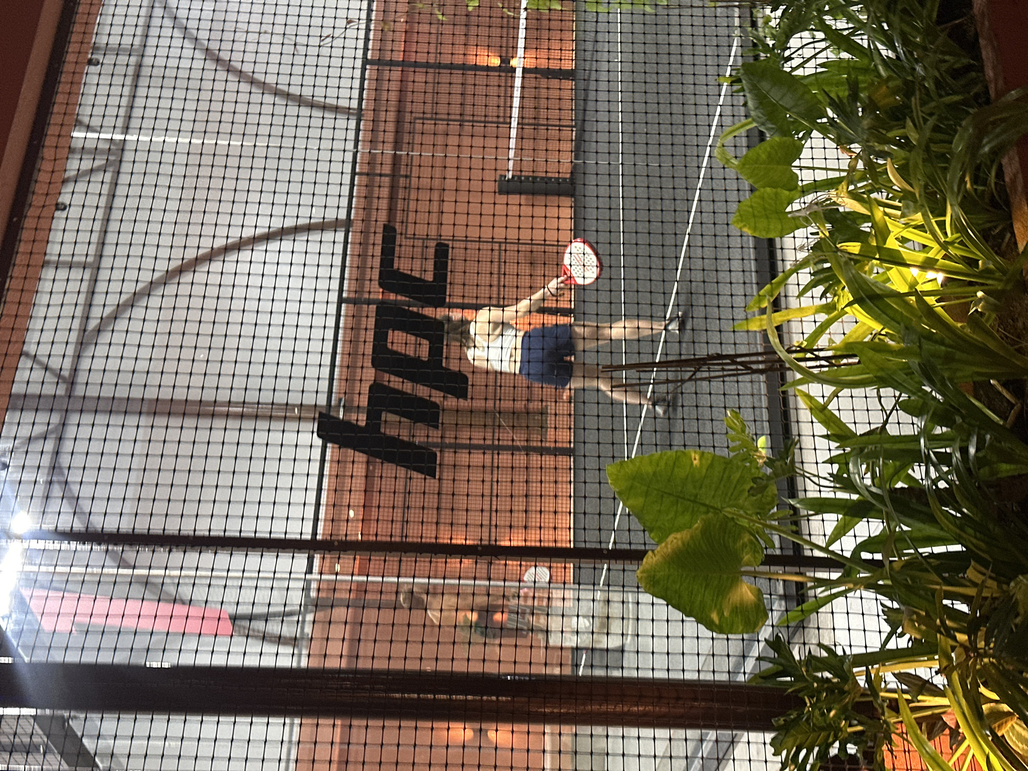 An overhead view shows a person playing padel on an indoor court in North Kuta, Indonesia. The court has a gray surface with white lines and a net dividing it. The player, wearing a white tank top and navy blue shorts, is in motion, holding a red and white padel racket. Shadows from the overhead netting create a grid pattern across the court. Large green plants with broad leaves are visible to the right of the frame, illuminated by warm lighting, suggesting an indoor setting with ambient light. The time of day appears to be evening or night due to the artificial lighting. Prominent black lettering, partially visible, spells out "HAPC" on the court's surface. The scene conveys a sense of active recreation.