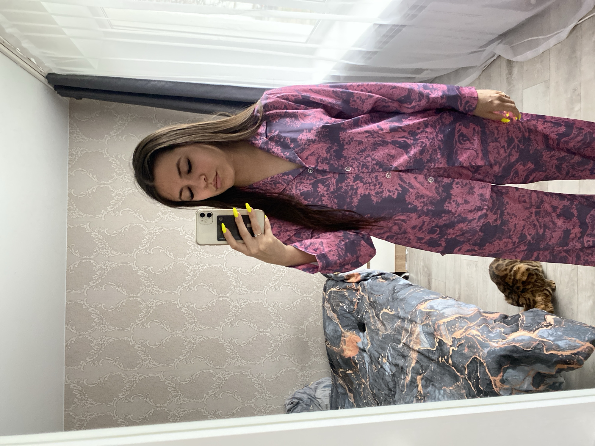 A woman is taking a selfie in a mirror. She is wearing purple patterned pajamas. The background includes patterned wallpaper, a window with white curtains, a bed with a patterned cover, and a cat. The location is possibly in Chaikovsky, Russia.