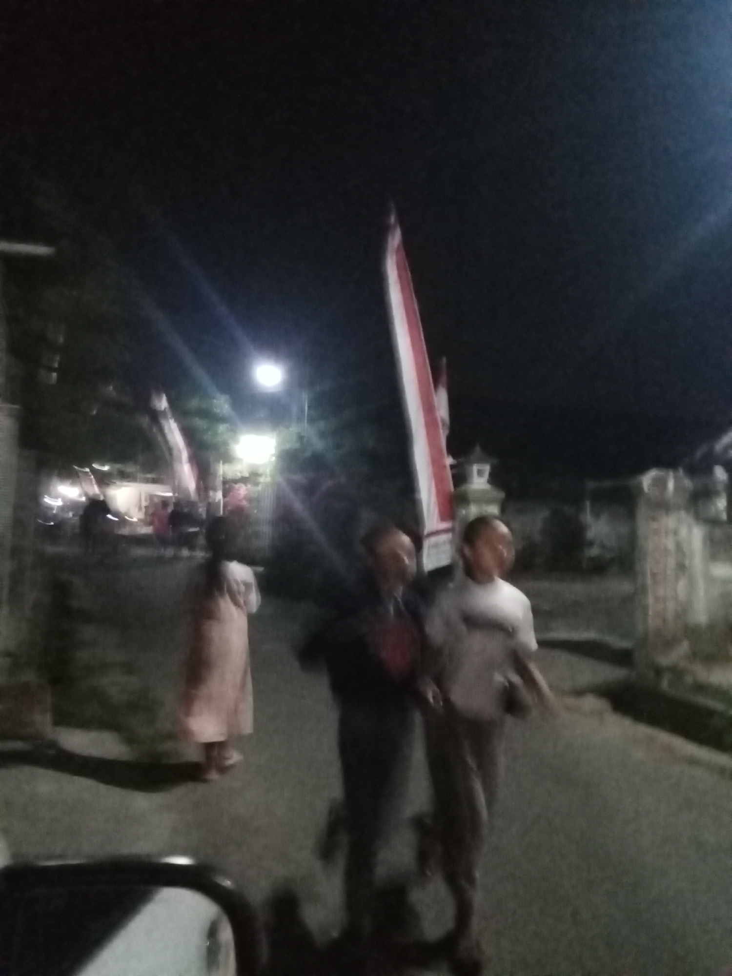 The image, taken at night in Penggaron, Indonesia, depicts a street scene with several individuals and a prominently displayed Indonesian flag. Two men are walking together in the foreground; one is wearing a dark shirt, and the other a white t-shirt. Behind them, a person in a light-colored dress is walking away from the camera. A tall Indonesian flag, red and white, is erected on the left side of the street, with another flag visible further in the background. Streetlights illuminate the scene, casting shadows and creating a somewhat blurry effect due to the low light conditions and possible motion blur. Buildings and foliage are discernible in the background, suggesting a residential or semi-urban environment. The overall atmosphere is that of a community gathering or procession during an evening event.
