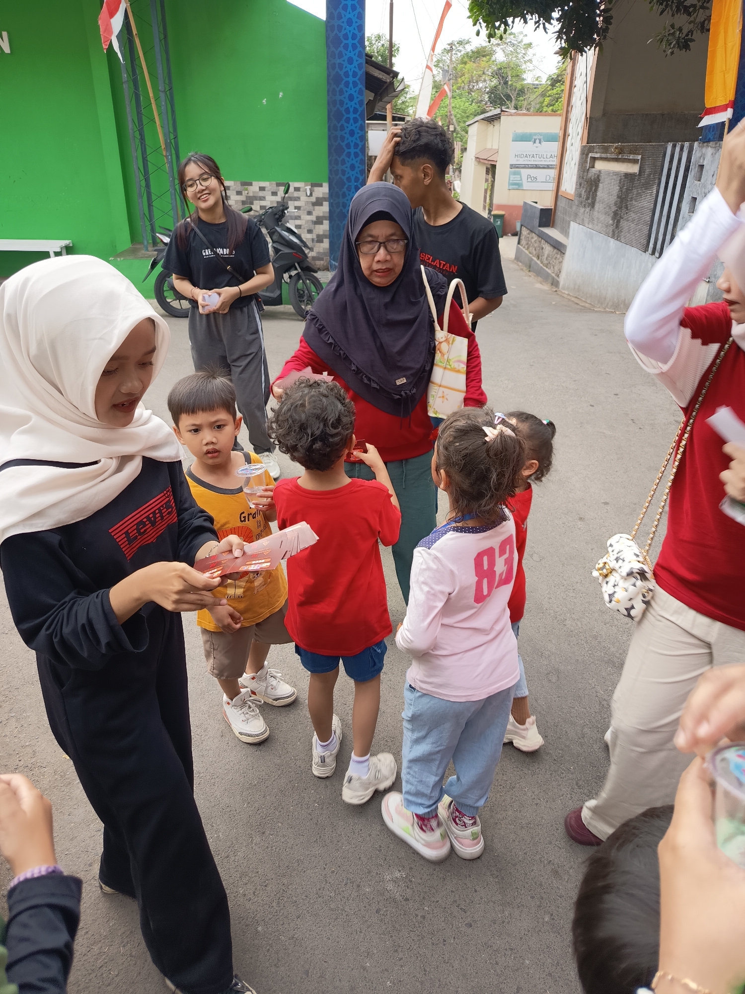 In Ngaglik, Indonesia, a group of people, including children and adults, are gathered outdoors on a paved surface. A woman wearing a white hijab and black top, holding a stack of red rectangular objects, is interacting with a young boy in a yellow shirt. Beside them, another child in a red shirt and blue shorts looks on, as does a young girl in a pink long-sleeved shirt with the number "83" on it. To the left, a woman in a black hijab and red top is also present. In the background, a motorcycle is parked near a green building. The scene appears to be a casual gathering, possibly an event or distribution, in a semi-urban environment.