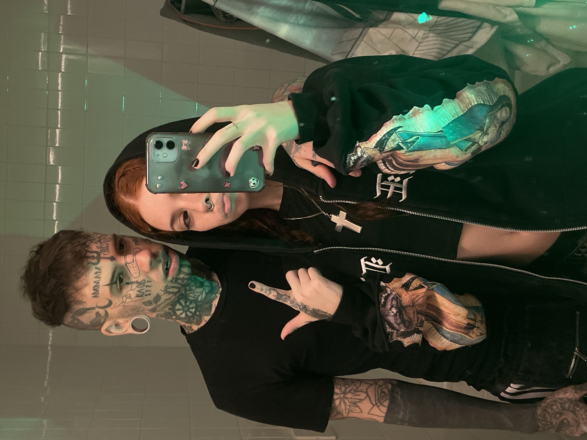 A couple is taking a selfie in a bathroom. The woman is holding a phone in her left hand and is wearing a black hoodie with a religious image on the sleeves and a cross necklace. The man has extensive face tattoos, stretched earlobes, and is wearing a black T-shirt. The bathroom has white tile walls and possibly a mirror reflecting the scene. There is no evidence in the image to indicate the location is Buenos Aires, Argentina.