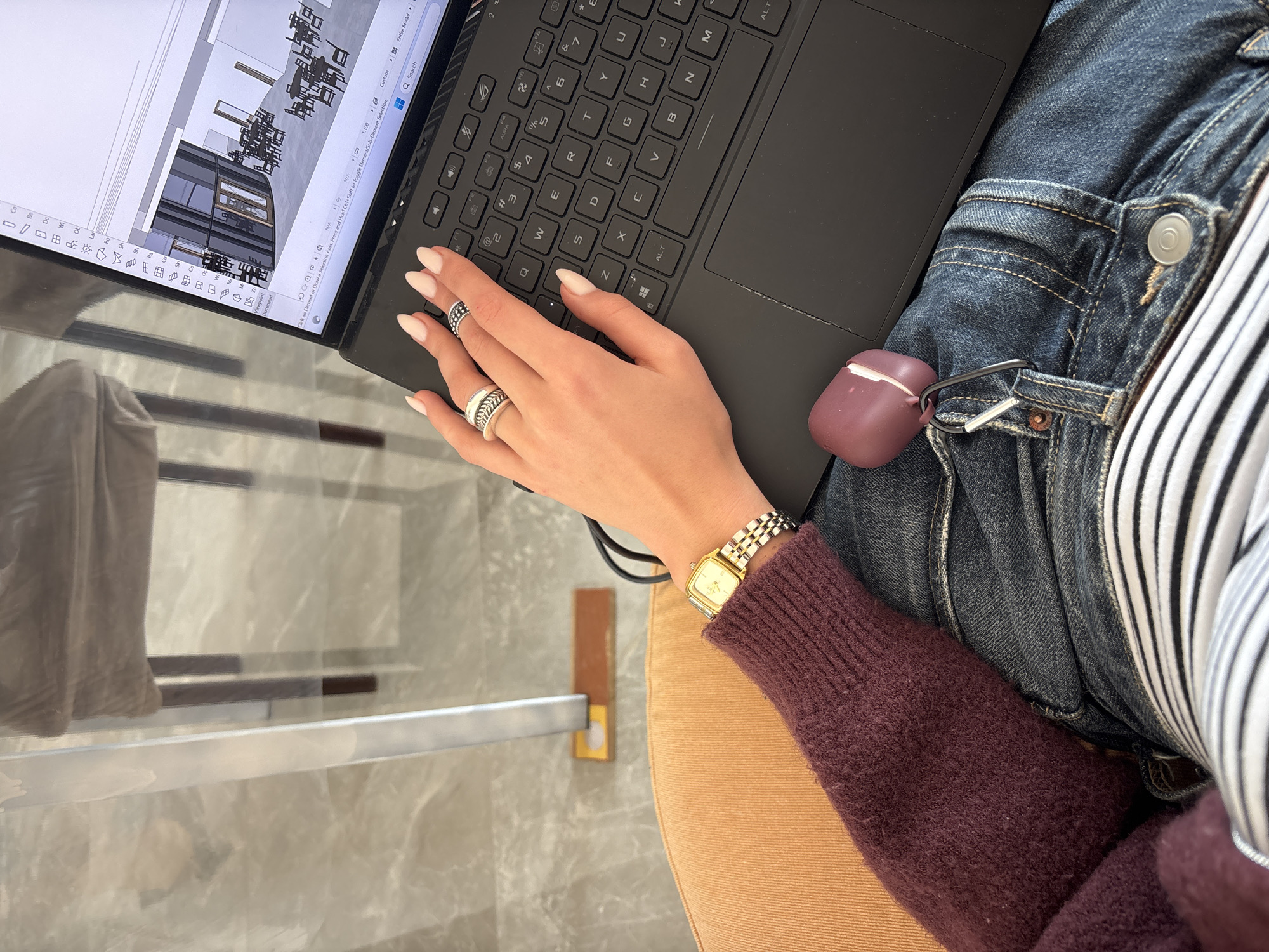 Here's a detailed description of the image:
**Scene:** An overhead shot of a person sitting at a glass table working on a laptop. The laptop screen displays a 3D architectural model.
**People/Subjects:** The image focuses on the person's hand on the keyboard, adorned with rings. The person is wearing a maroon sweater, a striped shirt underneath, and jeans.
**Objects:**
* Laptop
* Rings
* Watch
* Airpods case
* Jeans
* Sweater
* Striped Shirt
* Glass Table
* Chair
**Actions:** The person is using the laptop.
**Environment:** The background shows part of a room with neutral tones and simple furniture. The location in Abovyan, Armenia, is not discernible from the visual information provided.