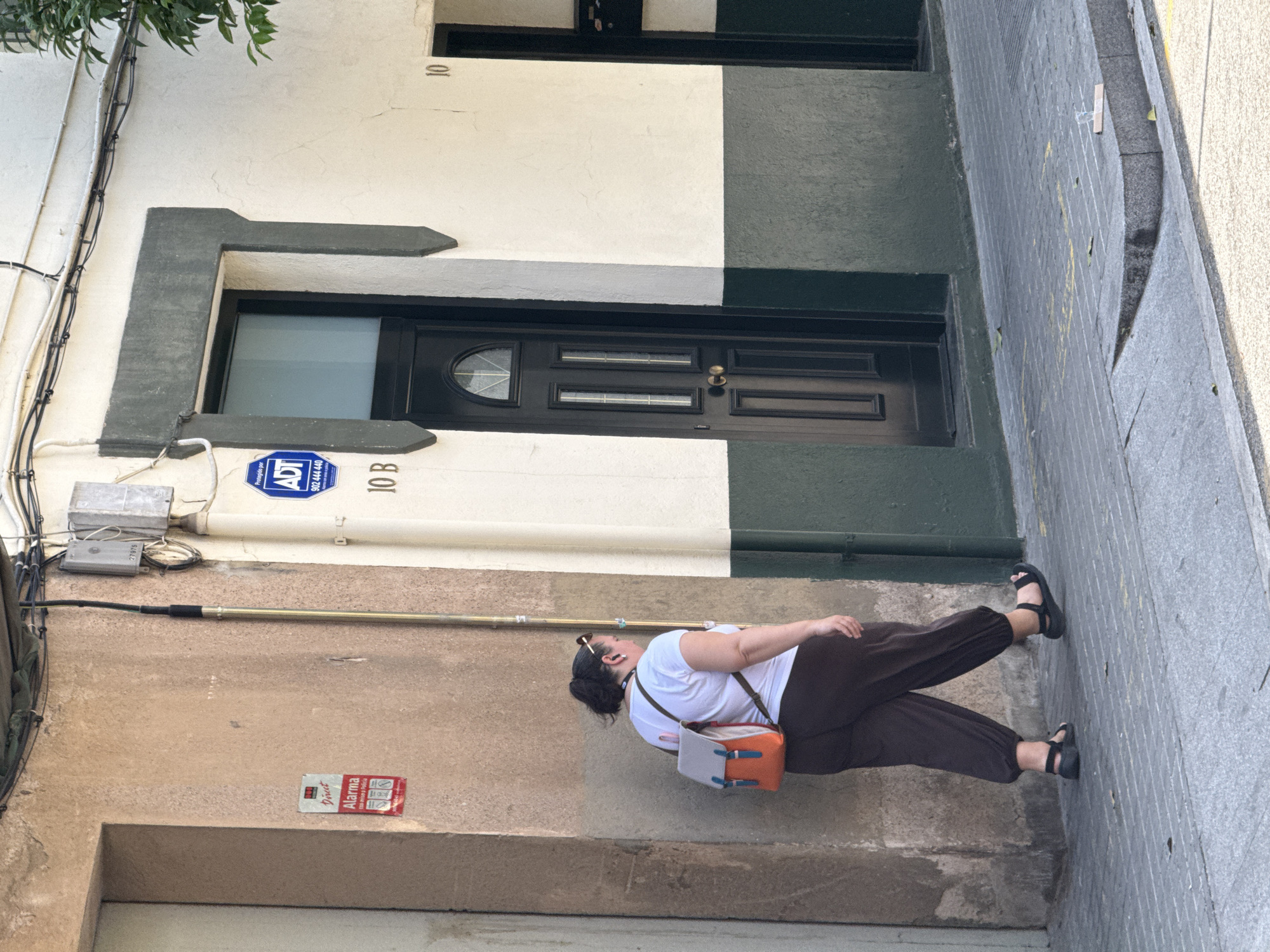 The image depicts a street scene in L'hospitalet De Llobregat, Spain. A woman, viewed from behind, is walking away from the camera on a sidewalk. She is wearing a white t-shirt, dark loose-fitting pants, and black sandals. She has a backpack with an orange and gray front pocket and brown straps on her back. She is also wearing sunglasses on the back of her head and has an earpiece in her left ear.
The woman is walking past a building with a textured, light brown wall on the left, which has a red and white alarm sticker. Adjacent to this is a building with a two-toned facade, split horizontally between white on the top and dark green on the bottom. This building has a dark-colored door with a semicircular window at the top and multiple rectangular panels. Above the door, there is an ADT security sign with a phone number. Further to the right, the building continues with more windows and what appears to be another entrance, also painted dark green on the lower half. The sidewalk is paved with gray square tiles and a ramped section made of darker granite. Scattered on the sidewalk are a few fallen leaves and some yellow markings. The overall lighting suggests a bright, sunny day.