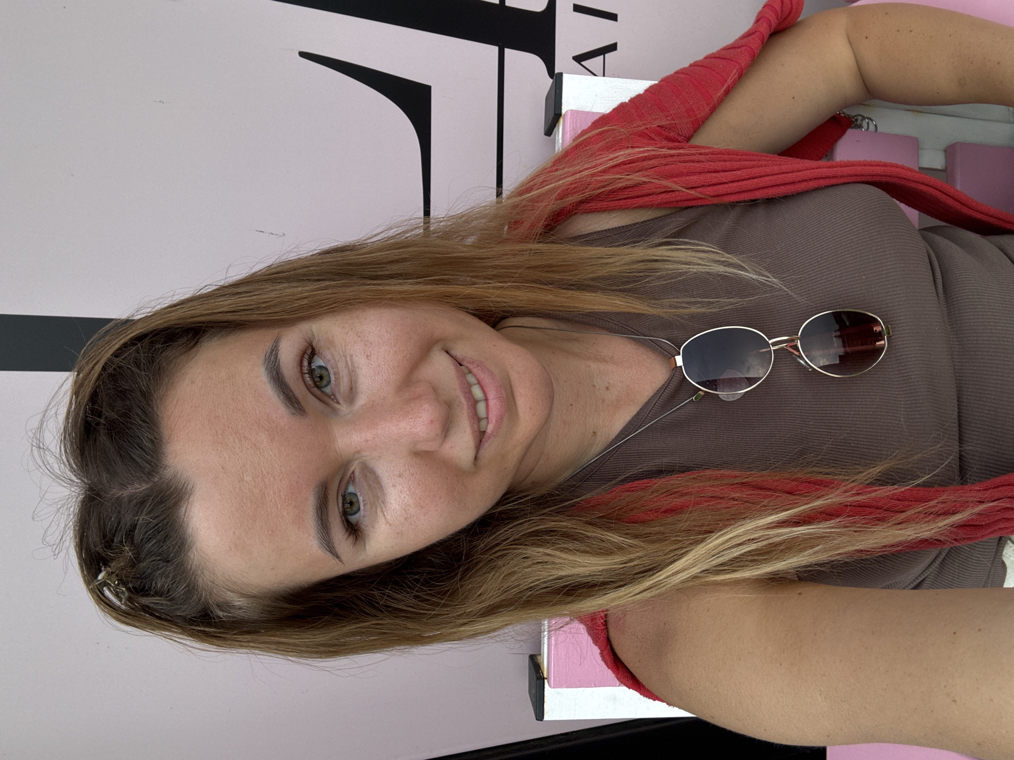 A medium shot shows a woman from the chest up, posing for a selfie outdoors. She has long, wavy brown hair with blonde highlights and a few strands are braided at the crown. Her eyes are green, and she has a light dusting of freckles across her nose and cheeks. She is wearing a brown, ribbed tank top with a red cardigan draped over her shoulders. A pair of gold-rimmed sunglasses with dark lenses hang from a silver chain around her neck. The background features a pale pink wall with a large black graphic. The woman is smiling slightly, looking directly at the camera.