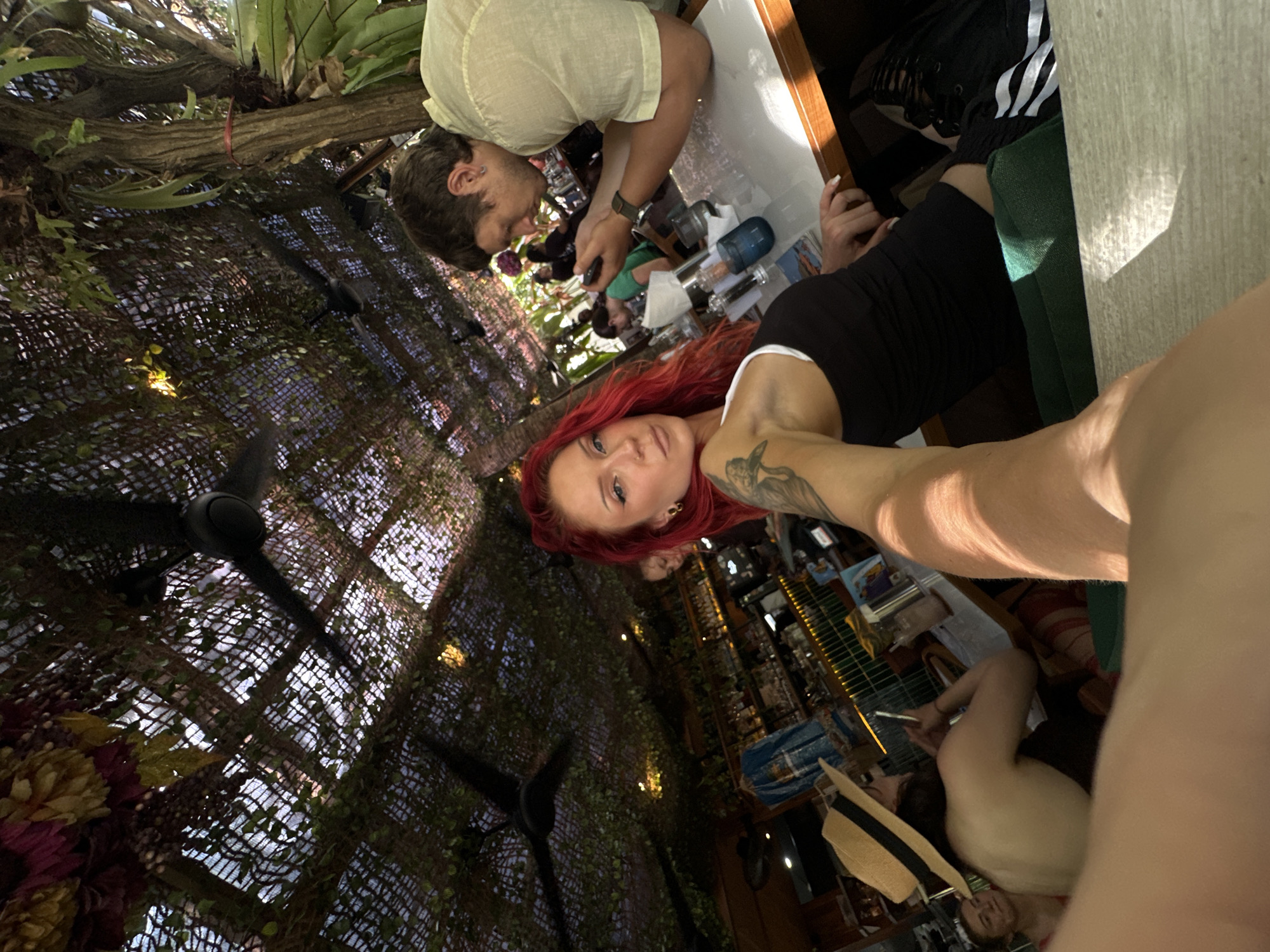 The image depicts an outdoor dining area, likely a restaurant or cafe, with a tropical ambiance. The ceiling is covered with a net-like material adorned with artificial foliage and some decorative flowers in burgundy and yellow. Two black ceiling fans are visible, indicating a warm climate. Sunlight filters through the canopy, creating dappled light effects.
In the foreground, a woman with vibrant red hair is positioned to the left, looking towards the camera. She is wearing a black tank top and has visible tattoos on her arm. To her right, a man is seated at a table, looking down at his phone, and wearing a light-colored short-sleeved shirt. The table is set with various items, including glasses, bottles, and possibly menus.
In the background, other patrons are seated at tables. One individual is wearing a straw hat. A bar area with shelves stocked with bottles is visible further back. The overall scene suggests a relaxed, social gathering in an open-air setting.
The location is identified as Mengwi, Indonesia, suggesting a Southeast Asian setting. The dense foliage, open-air structure, and likely warm weather are consistent with this region. The presence of ceiling fans further supports the tropical climate inference.