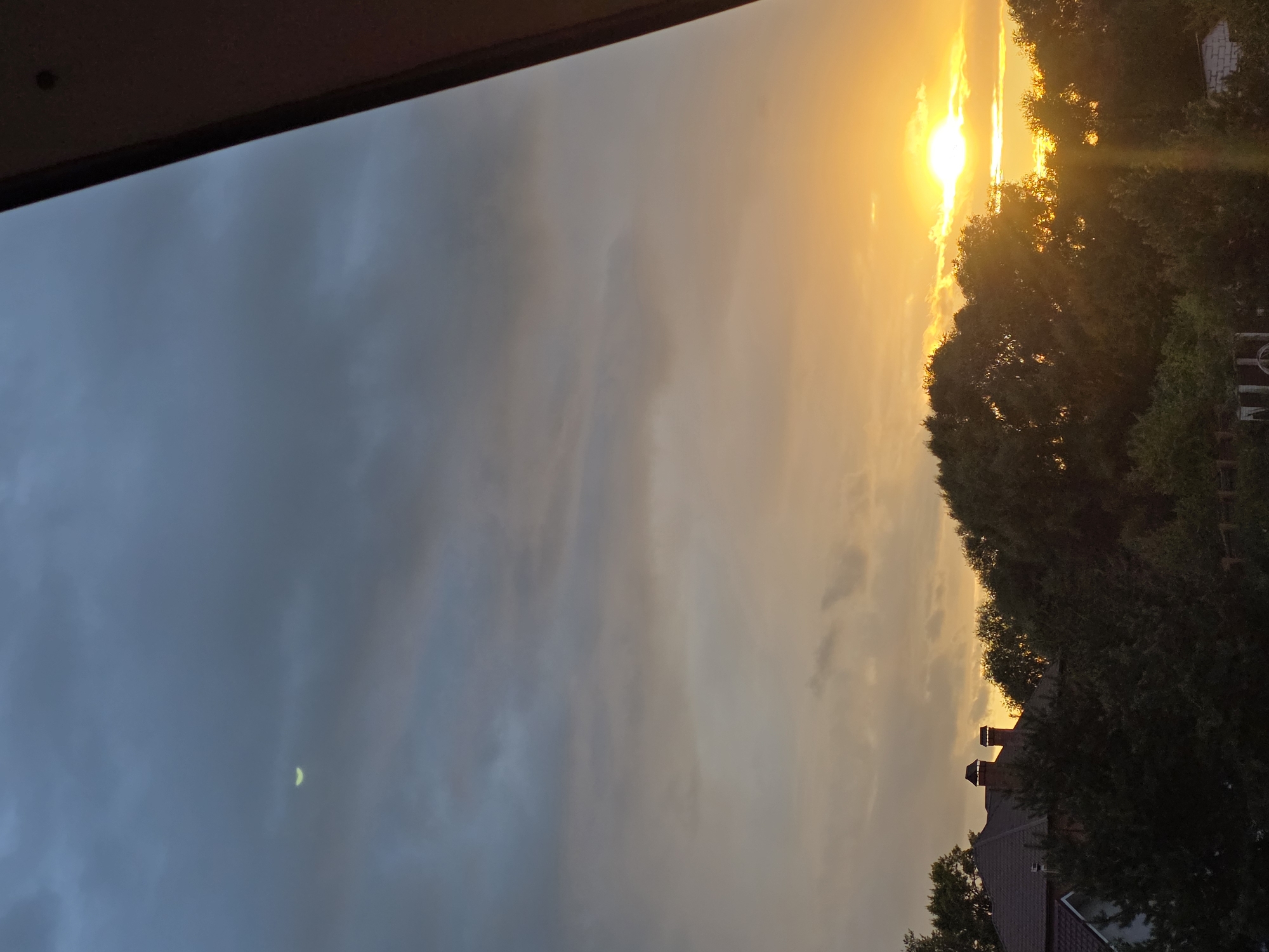 The image depicts a twilight sky over a residential area, possibly the city of Речицы, Russia. The sky is a gradient of colors, from a deep blue at the top to a warm orange and yellow near the horizon, indicating either sunrise or sunset. Clouds are present throughout the sky, with some appearing dark and heavy, suggesting the possibility of rain, while others are illuminated by the setting or rising sun.
In the upper left portion of the sky, a sliver of the moon is faintly visible. The horizon is dominated by the silhouettes of trees and rooftops. Several houses with dark-colored roofs and chimneys are visible amongst the trees. The trees are dense and appear to be deciduous, with full foliage.
The overall atmosphere is serene and natural, with a focus on the sky and the surrounding landscape. There are no people or animals visible in the image. The image appears to be taken from an elevated position, possibly a balcony or window, as suggested by the diagonal structure on the right side of the frame.