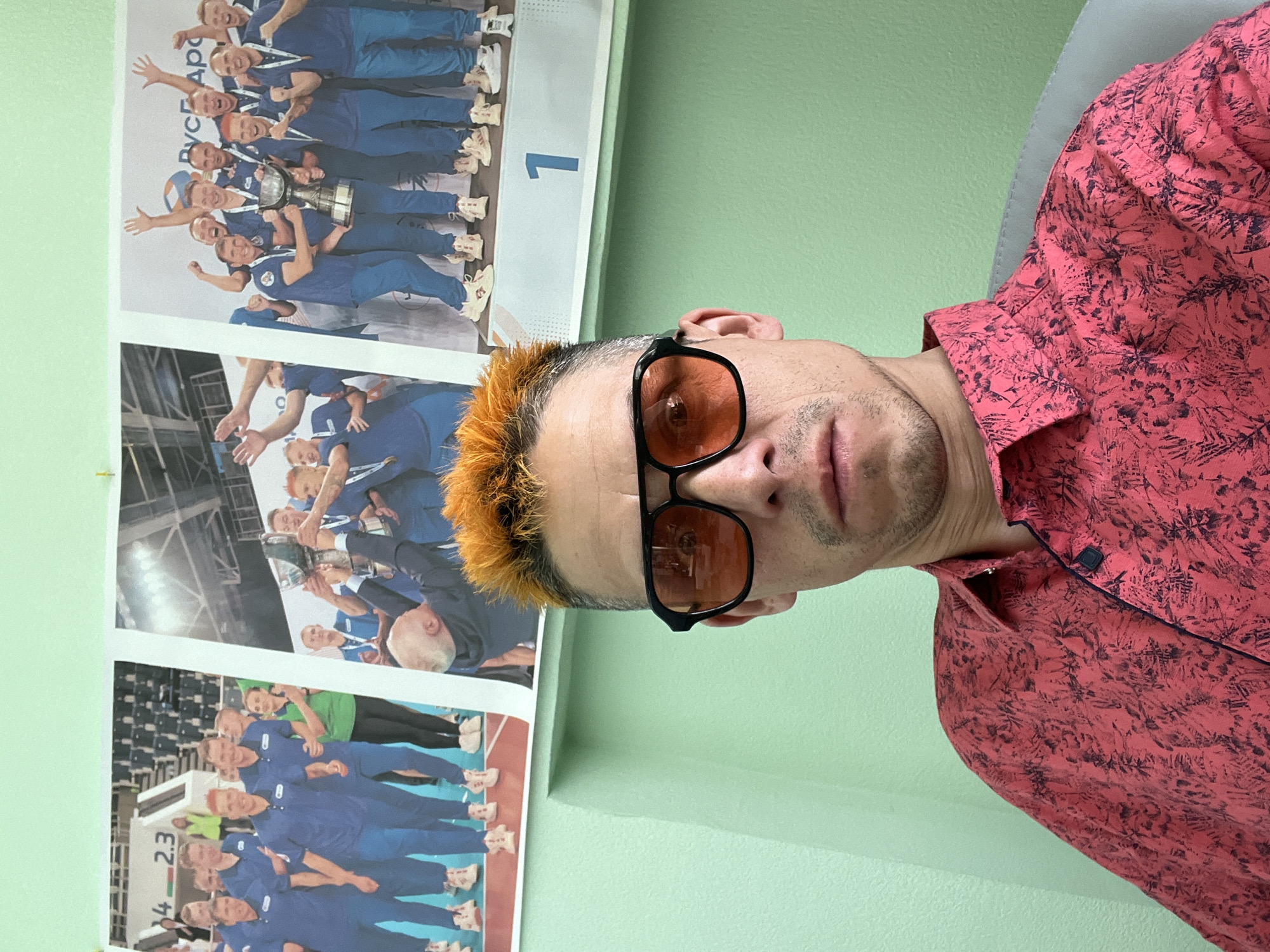 A man with orange-tipped hair and sunglasses is photographed indoors. He wears a pink patterned shirt and is positioned next to a light green wall. Two posters hang on the wall behind him: one displaying the number "1" and the words "РУСГард" above a team of people in blue uniforms, holding a trophy; the other below it shows another team, also in blue uniforms, with the number "4.23" visible. The location appears to be in Chaikovsky, Russia, based on the "РУСГард" text and the team's uniform colors and number signs on the team.