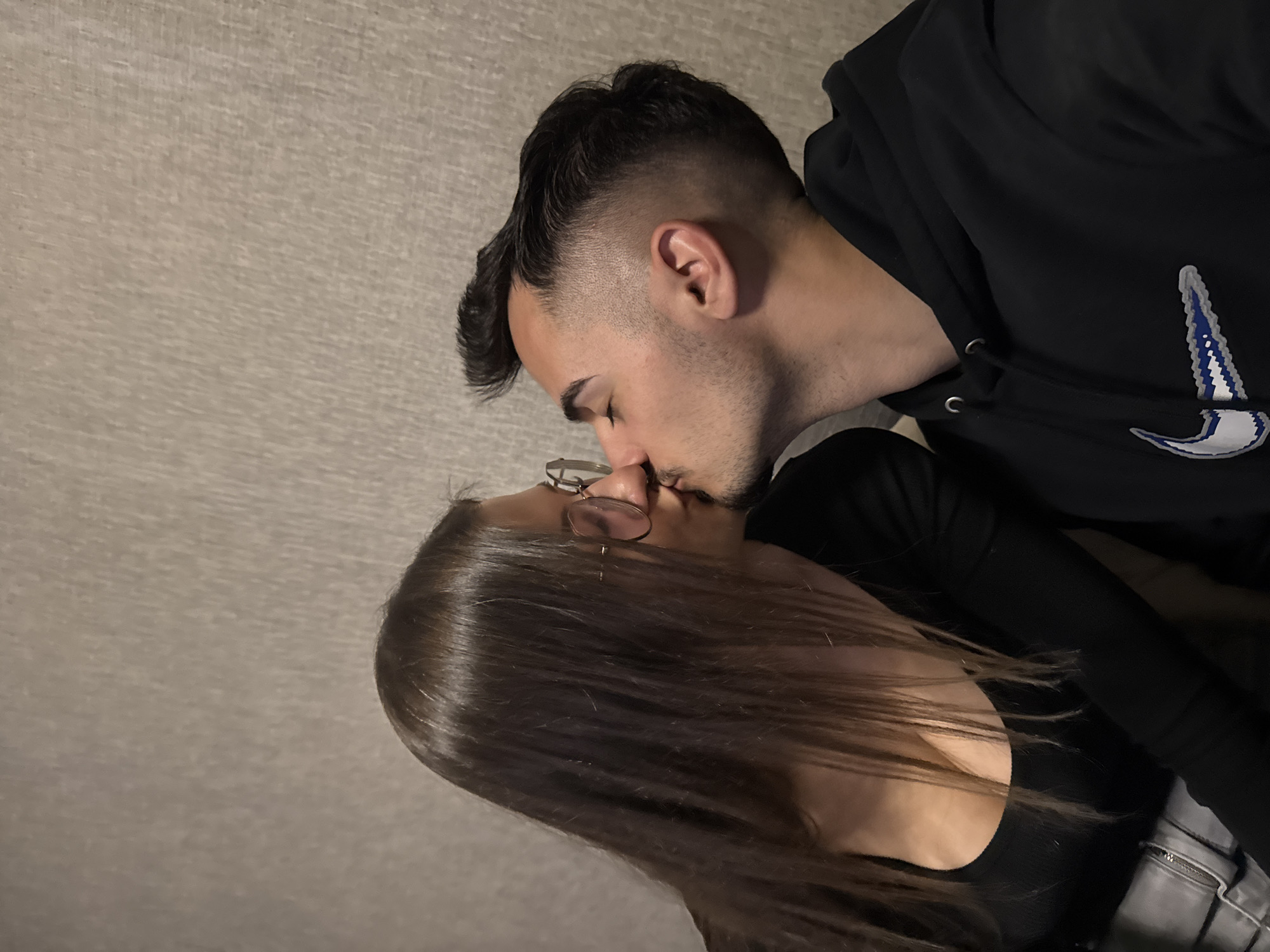 A couple is kissing indoors. The man has short dark hair, and a black hoodie with a white and blue logo. The woman has long brown hair and is wearing glasses and a black top. The background is a neutral color. The photo was not taken in Sofia, Bulgaria.