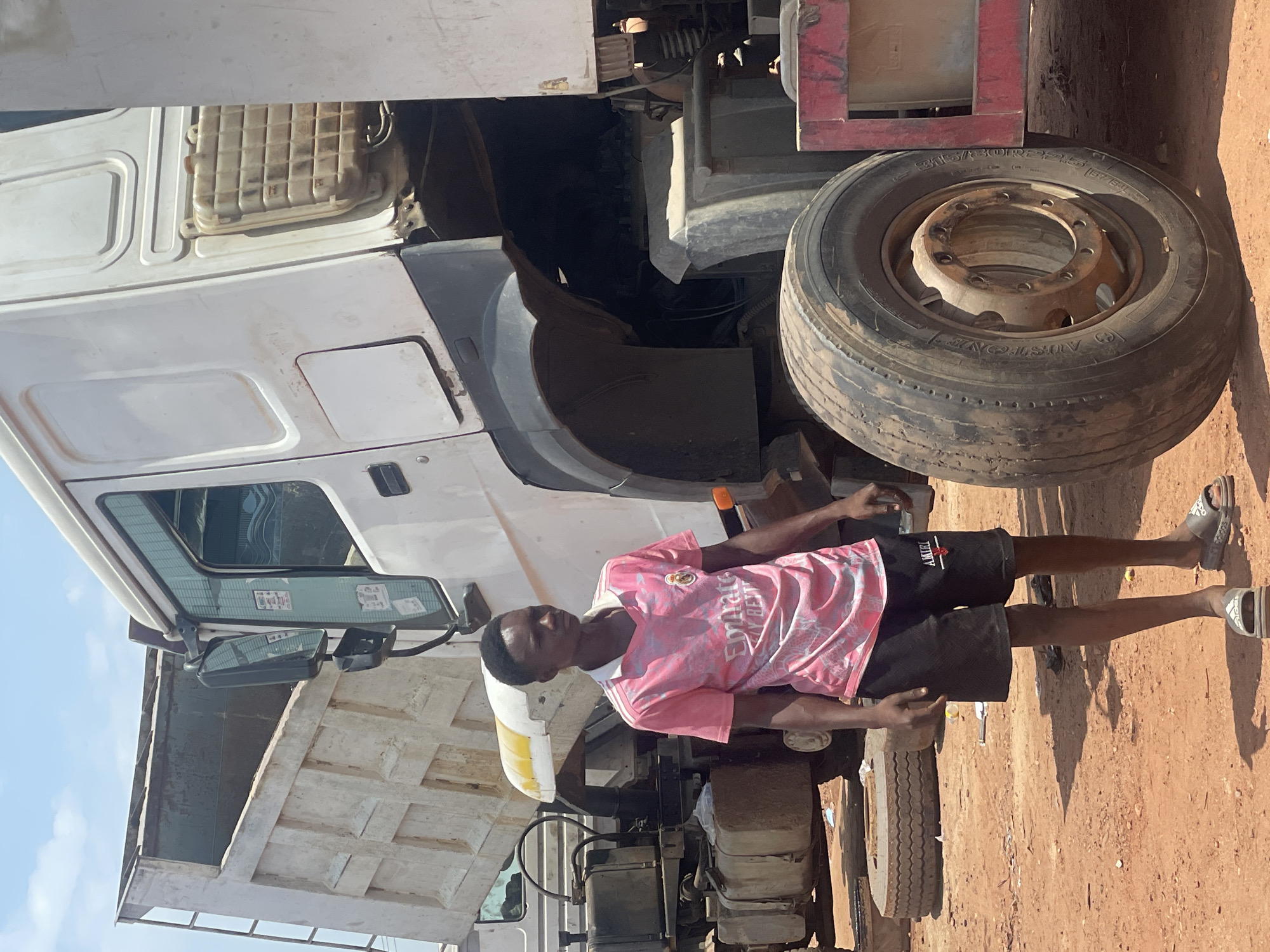 A young man stands near the front of a white dump truck in Kwali, Nigeria. The truck's cab is partially visible, as is one of its large front tires. The man wears a pink t-shirt and black shorts and gray sandals. The ground is brown dirt. The sky is blue.