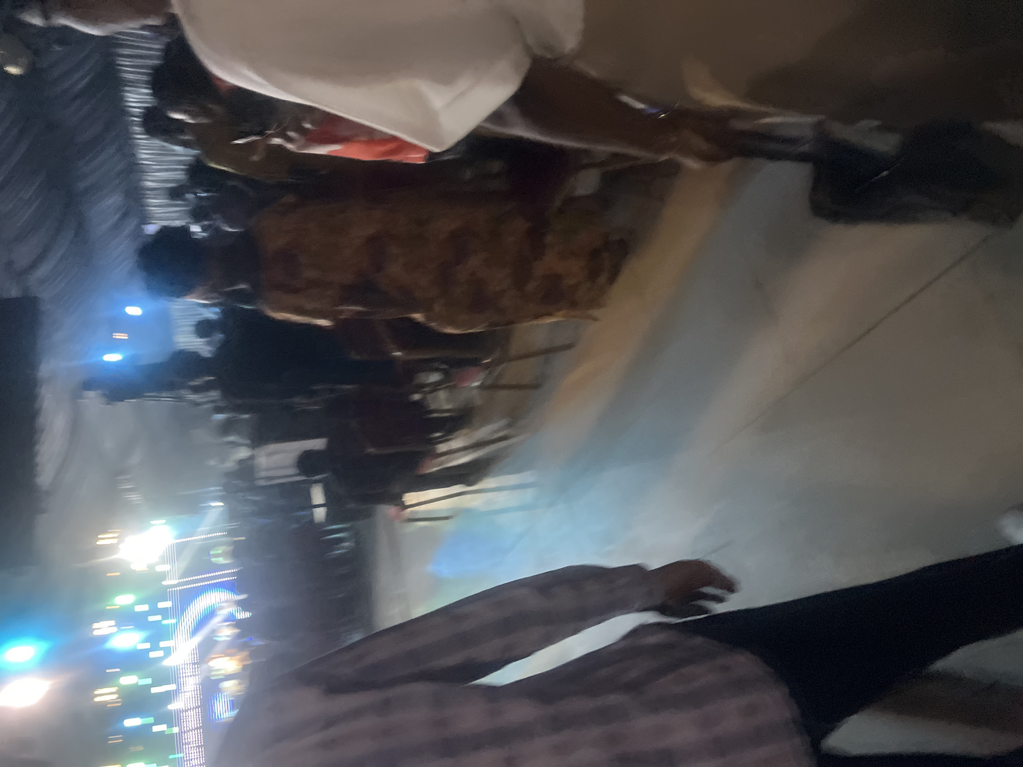 This image depicts an indoor event in Galadimawa Village, Nigeria, characterized by dim lighting and artificial illumination from a stage. The environment is a large, enclosed space with a dark, textured ceiling, possibly fabric, creating undulations. The floor is a light-colored, reflective surface, potentially polished concrete or tiling, which exhibits distinct reflections of the bright stage lights.
In the foreground, two individuals are partially visible, both facing away from the camera. On the left, a person is seen from the chest down, wearing a light-colored plaid long-sleeved shirt and dark trousers, stepping forward. On the right, another individual, visible from the torso down, is wearing a white top and light-colored lower garment, holding a dark, rectangular object (possibly a bag or folder) in their right hand.
The midground and background are occupied by a crowd of numerous individuals, most appearing to be standing or moving. Many are blurred due to low light and potential motion. A person in a brown patterned dress or top is prominent in the mid-right. Further back, several red-backed chairs are visible, suggesting designated seating areas within the venue.
At the far end of the room, a brightly lit stage or backdrop is present. This area is heavily illuminated by multiple vertical and horizontal light fixtures emitting vibrant blue, green, and white light, which create significant flares and streaks across the image, reflecting off the floor. The overall scene suggests a public gathering or performance event.