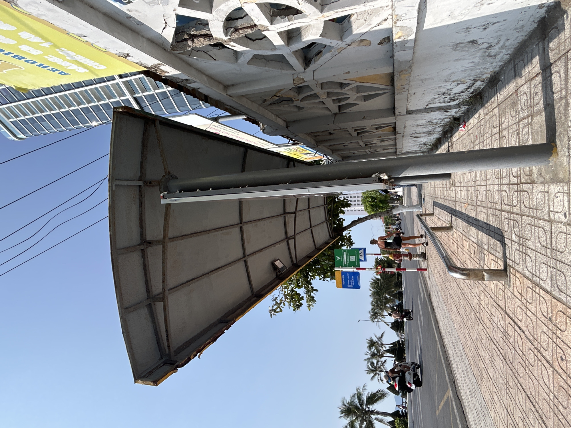 This image captures a bright daytime scene along a street in Bắc Nha Trang, Vietnam. The perspective is taken from beneath a wide, aged concrete overhang or awning, which displays decorative geometric and circular patterns on its underside. This structure shows signs of significant wear and tear, with cracks and crumbling concrete visible.
A yellow banner hangs from the overhang, partially visible with the words "AEROBI", "SINCE", "14h", and "6h" printed on it. Beyond the overhang, reflections of a modern glass-facade building are visible in the distance.
The foreground features a patterned sidewalk with square tiles, running parallel to a street. A silver metal railing is mounted along the sidewalk edge. Several individuals are present: one person is walking away on the sidewalk, another stands near a utility pole, and further down the street, multiple motorbikes with riders are visible, suggesting bustling activity.
The street is lined with numerous palm trees and other green foliage, indicative of a tropical climate. The sky is clear and a vibrant blue, with strong sunlight casting distinct shadows across the sidewalk and street, suggesting it is either morning or late afternoon. A small blue sign mounted on a pole further down the sidewalk clearly displays "Tên tuyến", which translates from Vietnamese as "Route Name". The overall scene portrays an active urban environment in a warm, sunny setting.